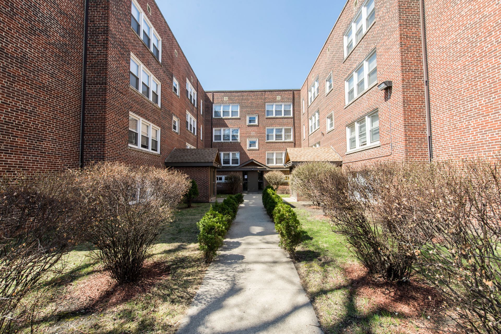 Apartment building entrance with brick facade, pathway, and landscaping.