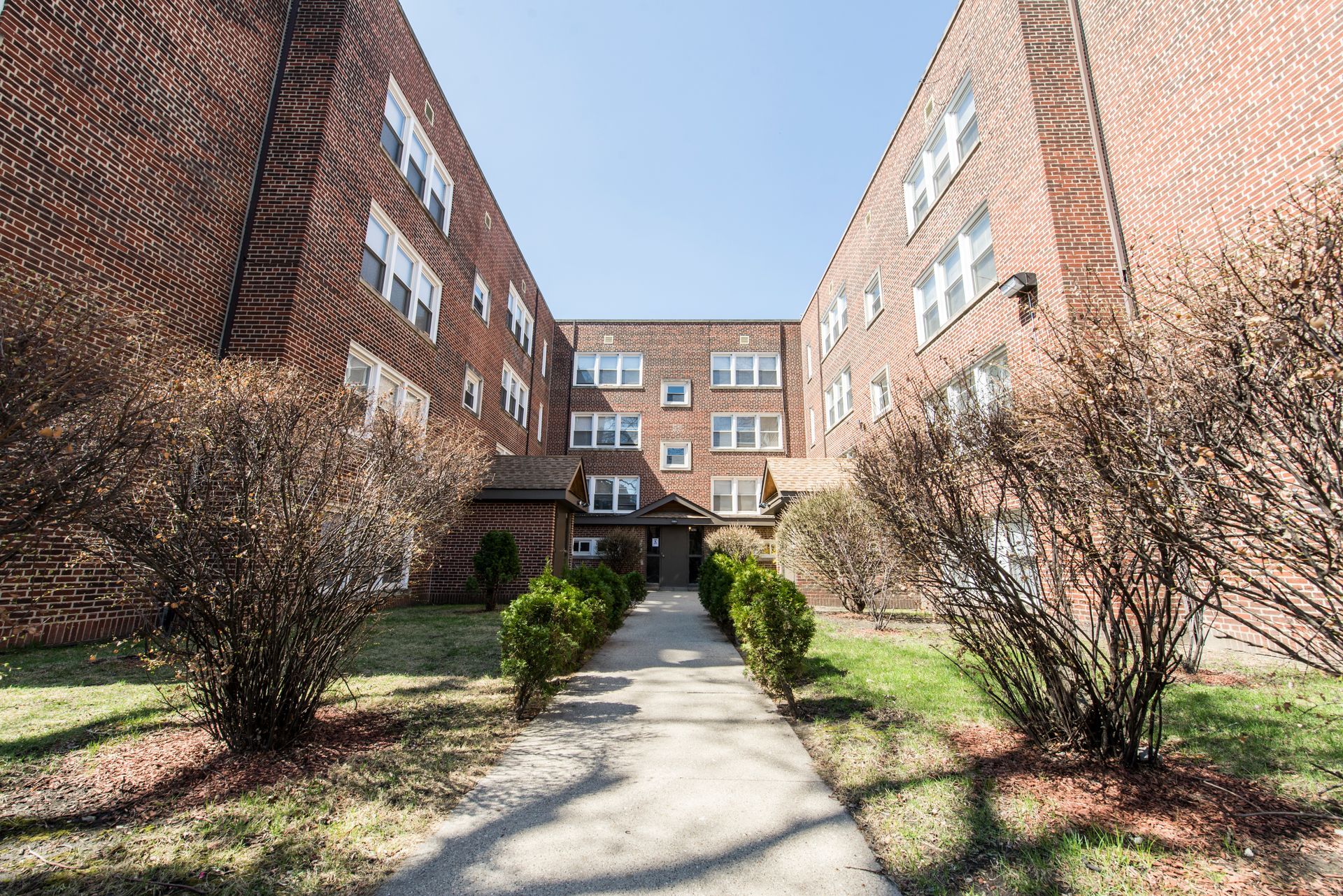 Brick apartment building with pathway leading to entrance, surrounded by bushes and grass.