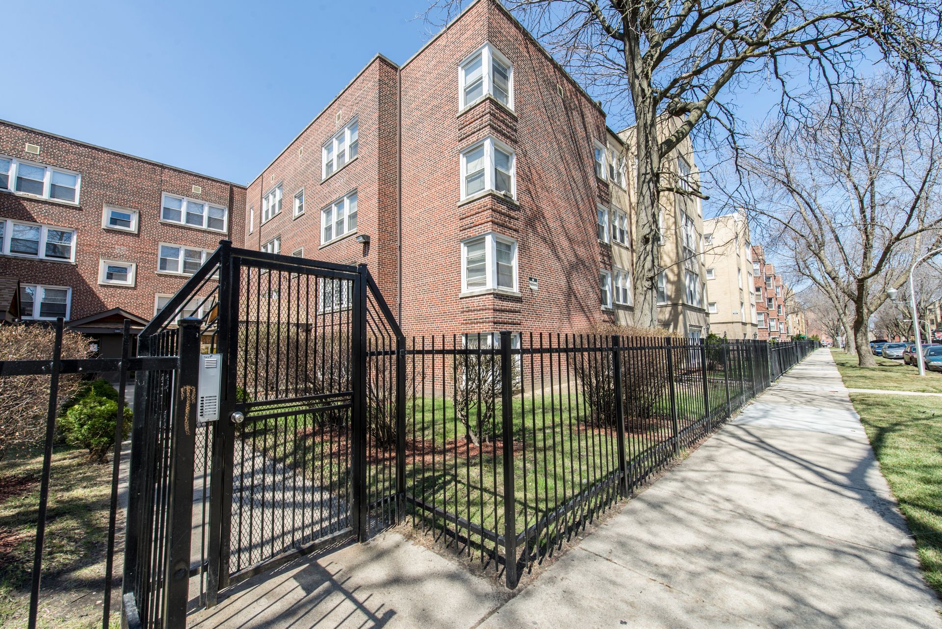 Brick apartment building behind black metal fence on sunny sidewalk.