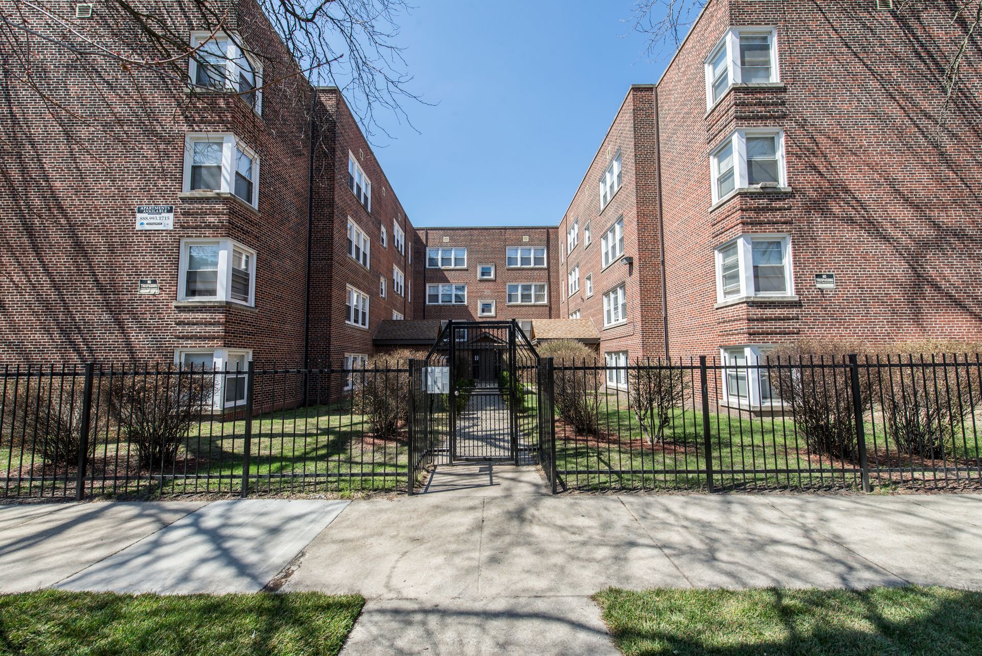 Brick apartment building with black fence, trees, and blue sky.
