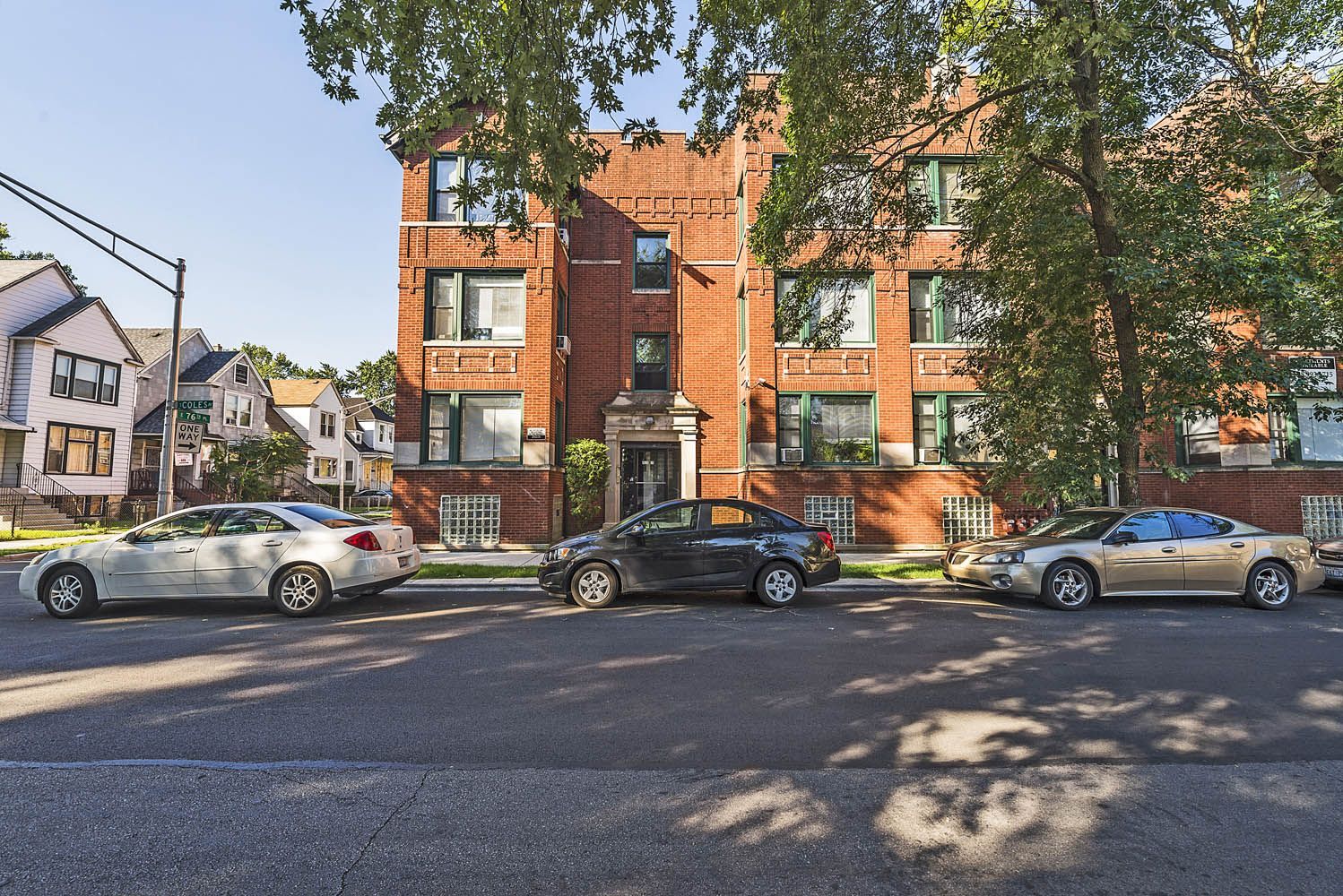 Brick apartment building with parked cars on a sunny street, trees provide shade.