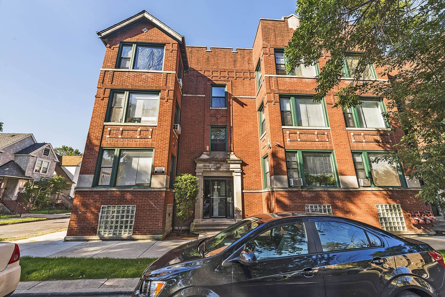 Red brick apartment building with cars parked out front, on a sunny day.