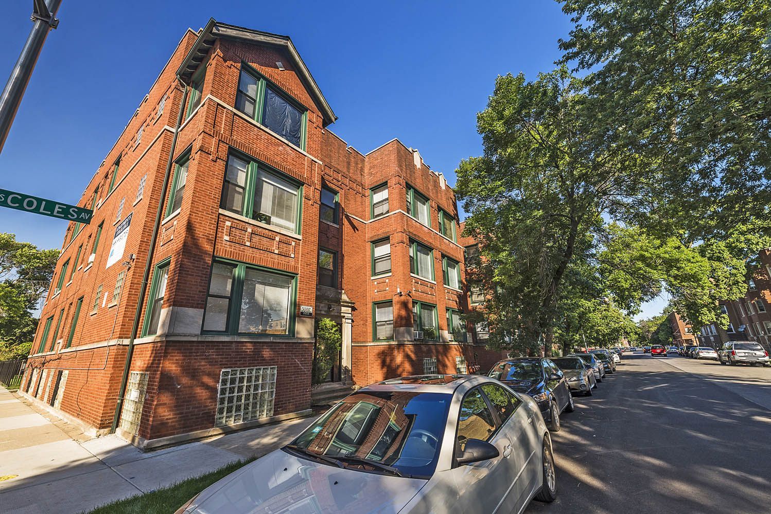 Red brick apartment building on a sunny street, parked cars, green trees, street sign visible.