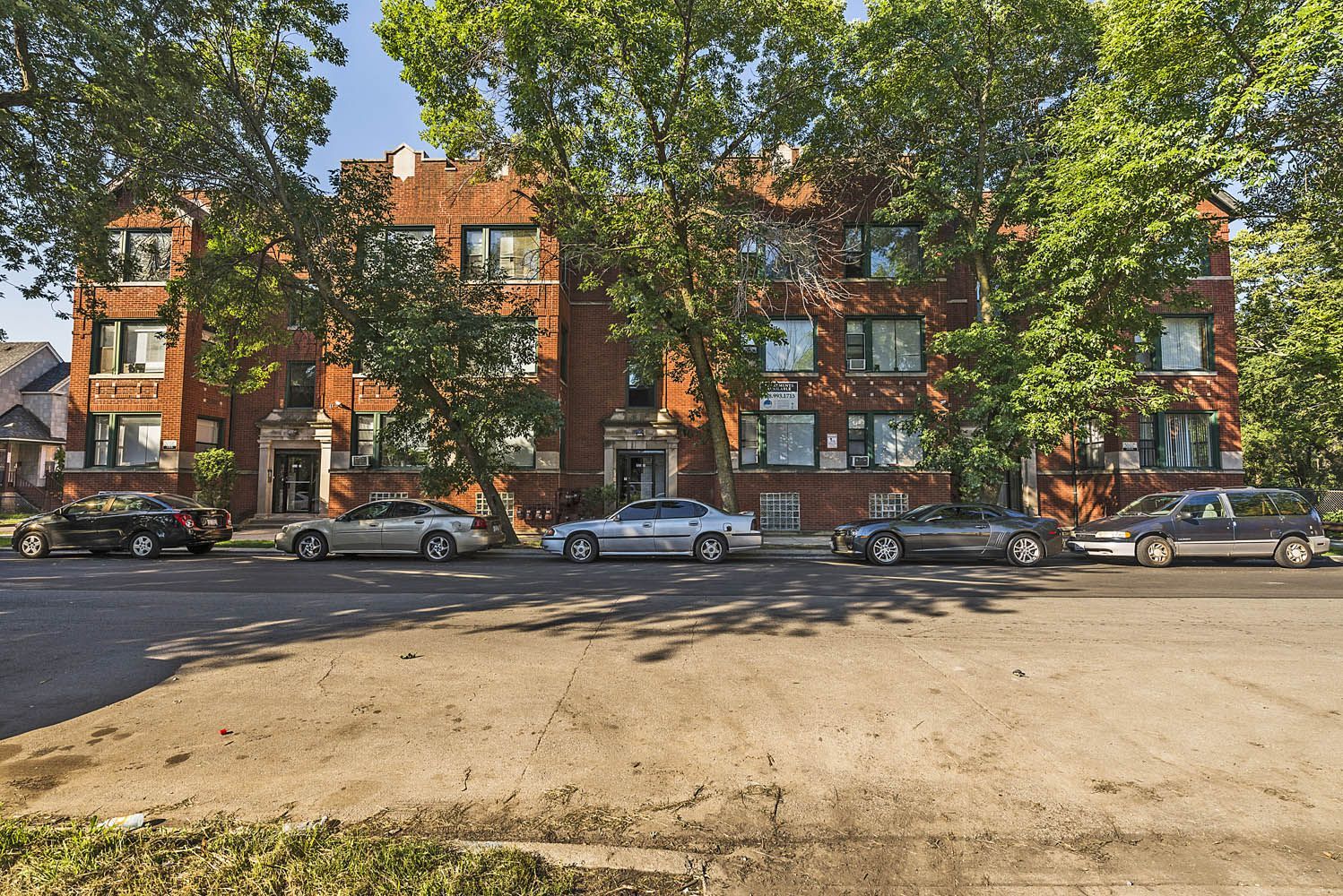 Red brick apartment building with parked cars along street, trees in front.