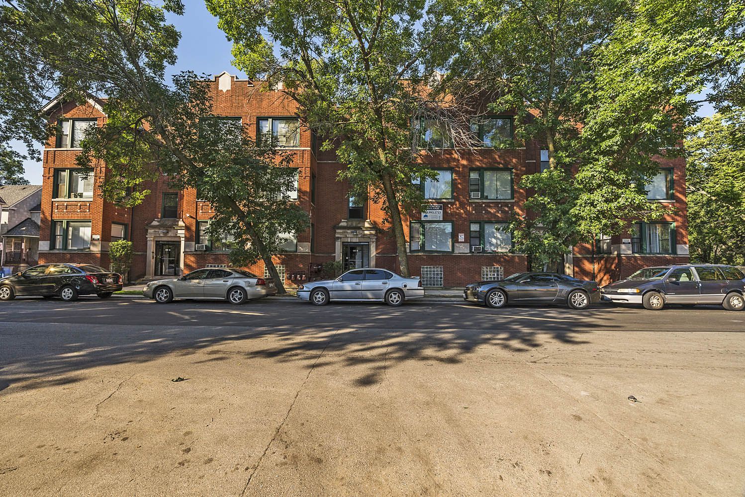 Brick apartment building with parked cars on a sunny day. Trees in front cast shadows.