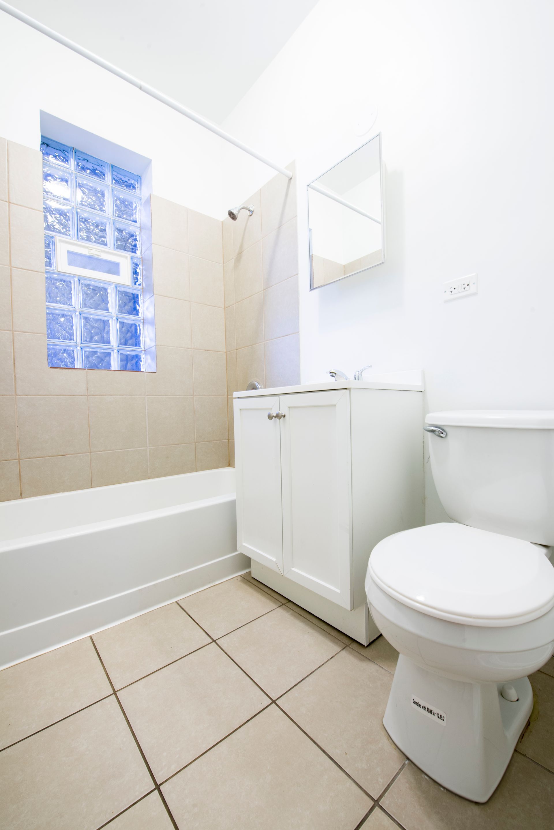 Bathroom with white toilet, vanity, and tan tile floor; window with blue glass blocks.