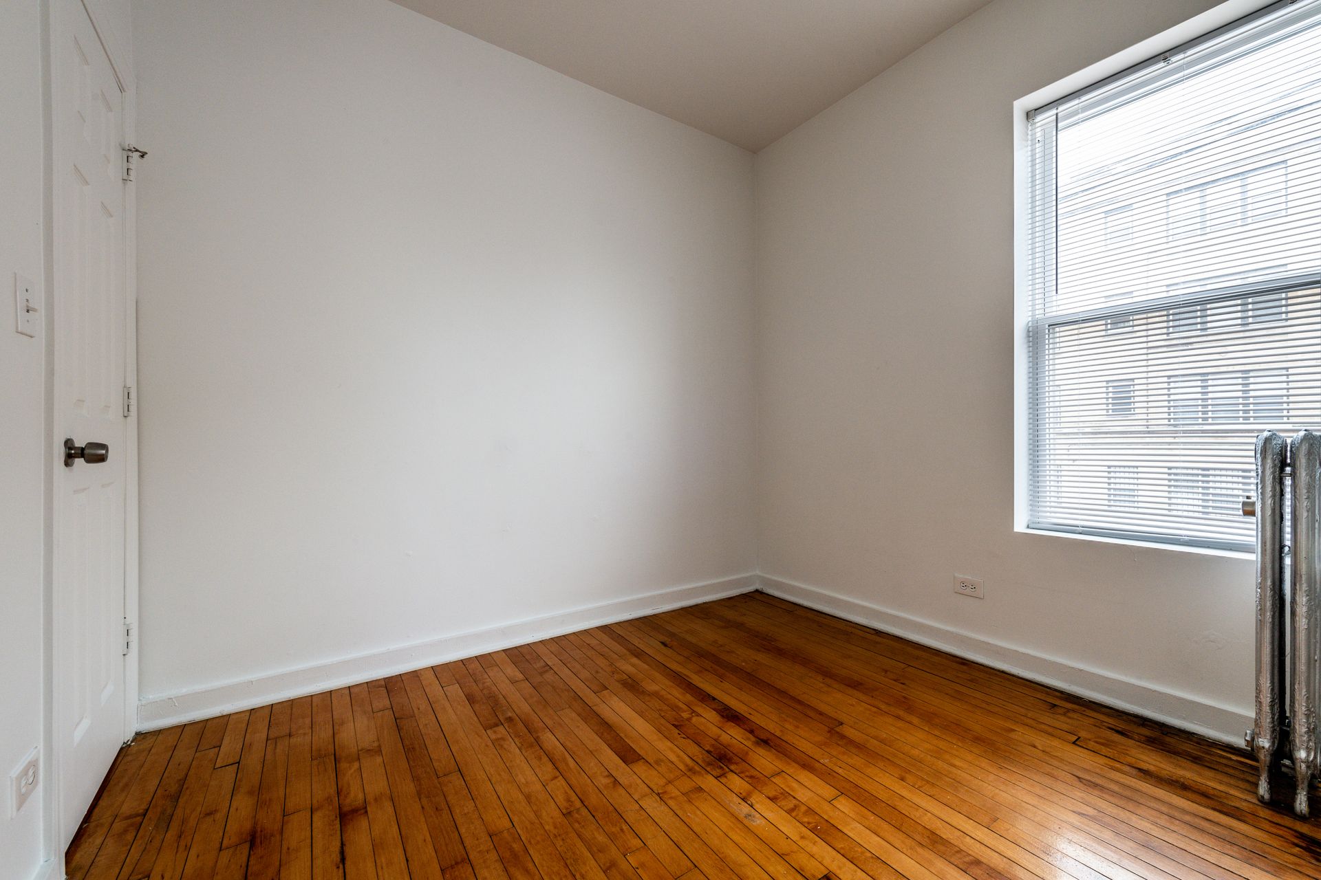 Empty room with wood floor, white walls, radiator, window with blinds, and a closed door.