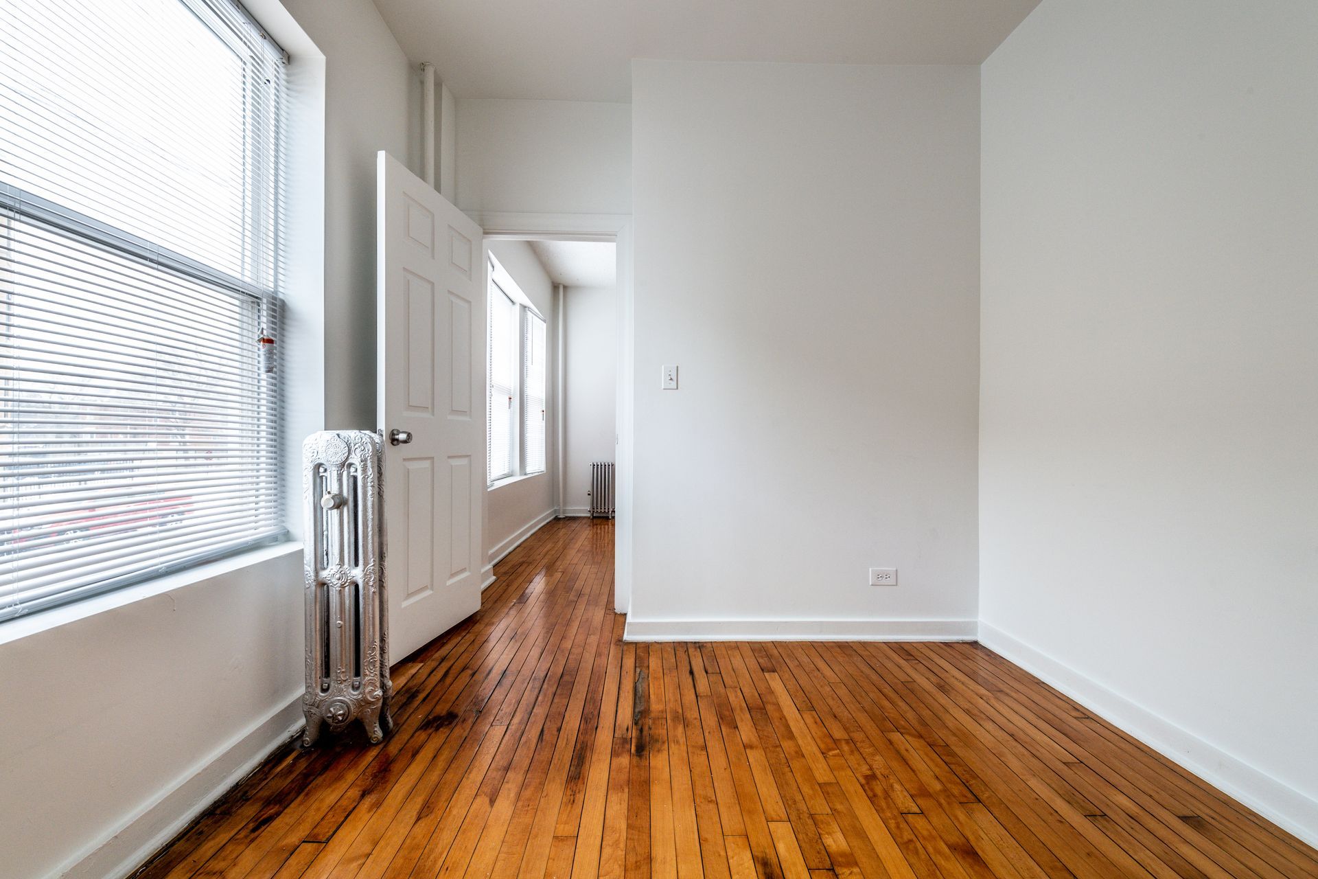 Empty room with wood floor, white walls, two windows with blinds, and open door.