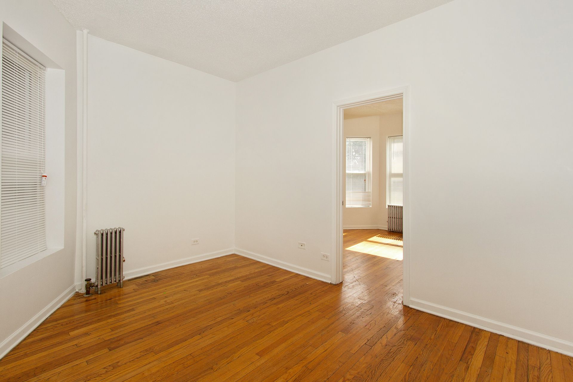 Empty room with hardwood floors, white walls, and a doorway leading to another room with windows.