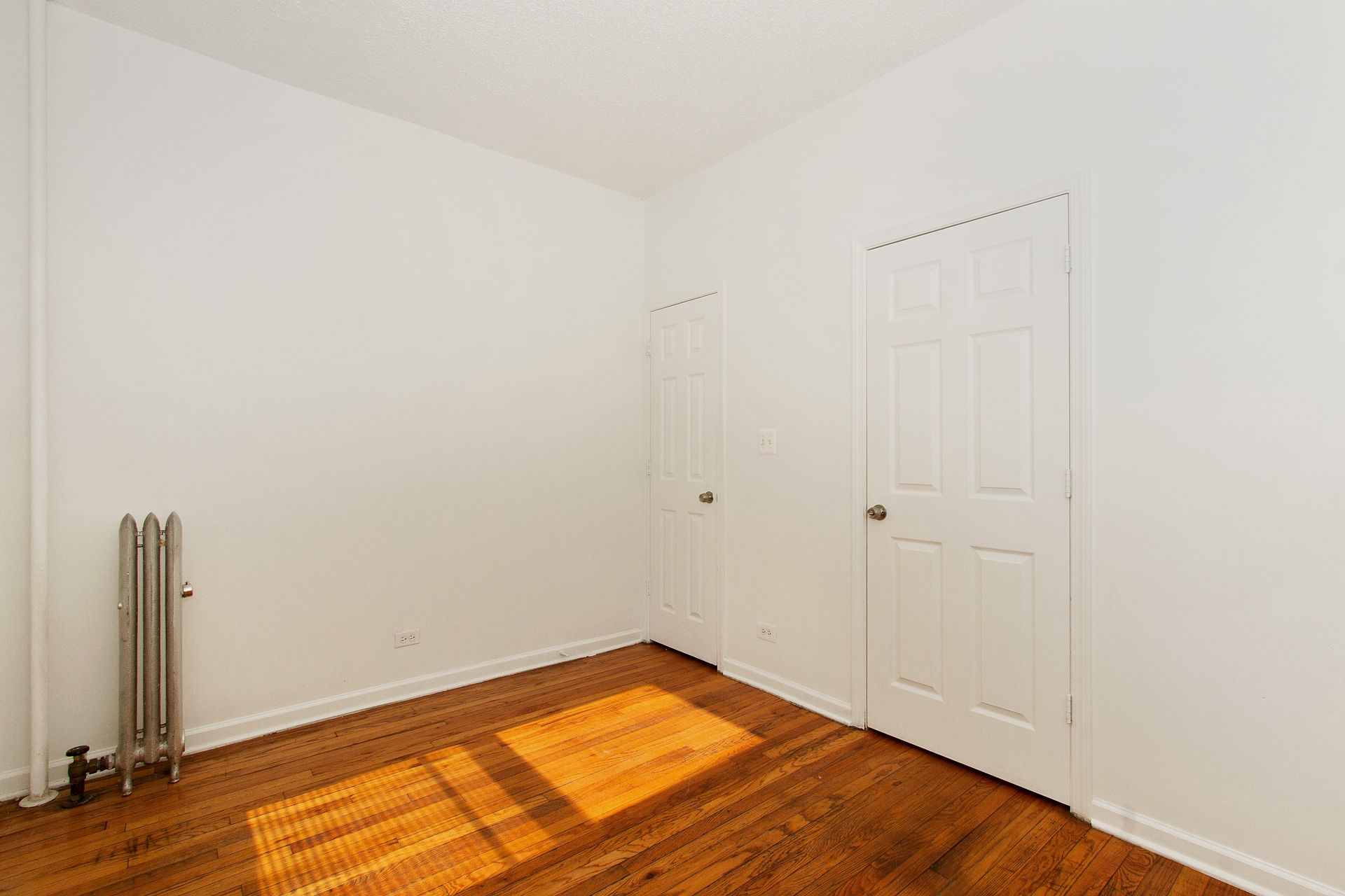 Empty white-walled room with hardwood floors, two doors, and a radiator. Sunlight streams in.