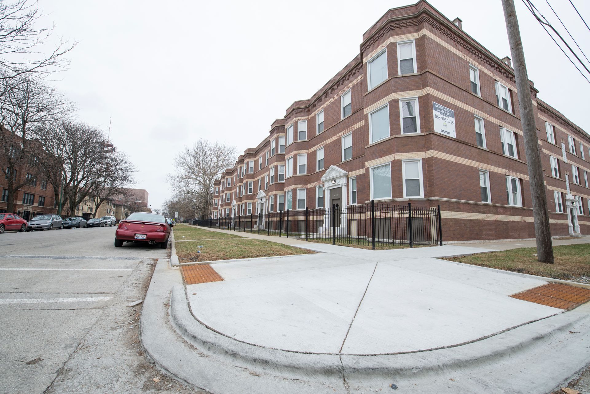 Brick apartment building on a street corner, gray sky, cars parked along the street.
