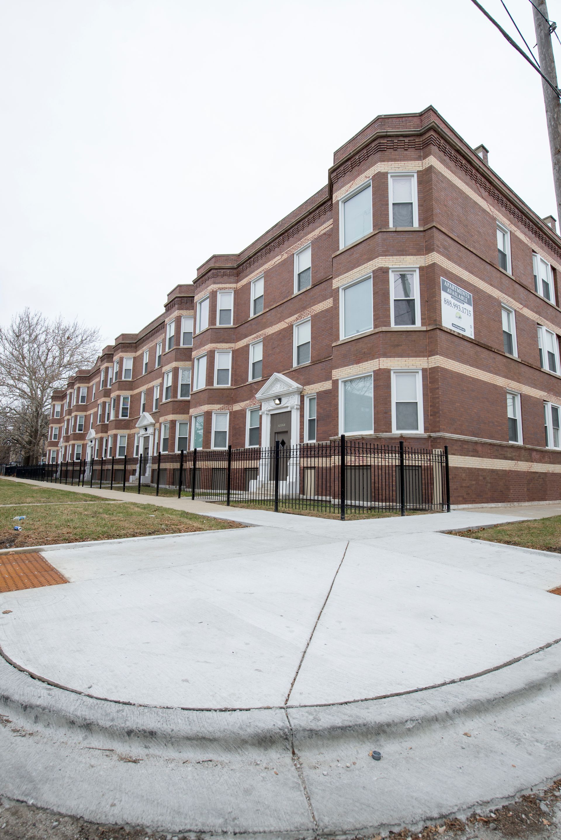 Brick apartment building with bay windows on a cloudy day. Sidewalk and fenced front yard.
