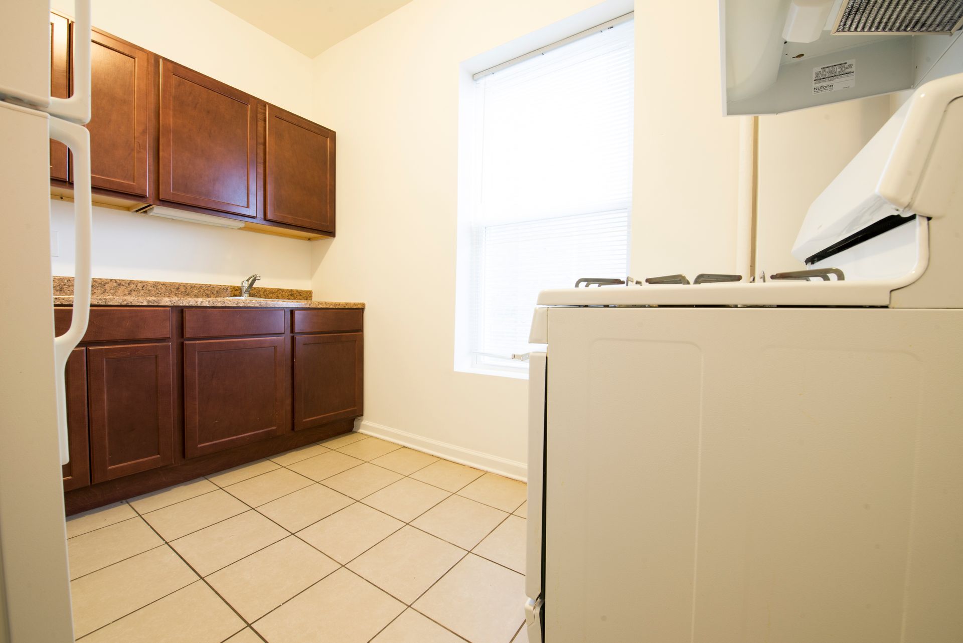 Kitchen with dark brown cabinets, beige tile floor, white appliances, and a window with a blind.