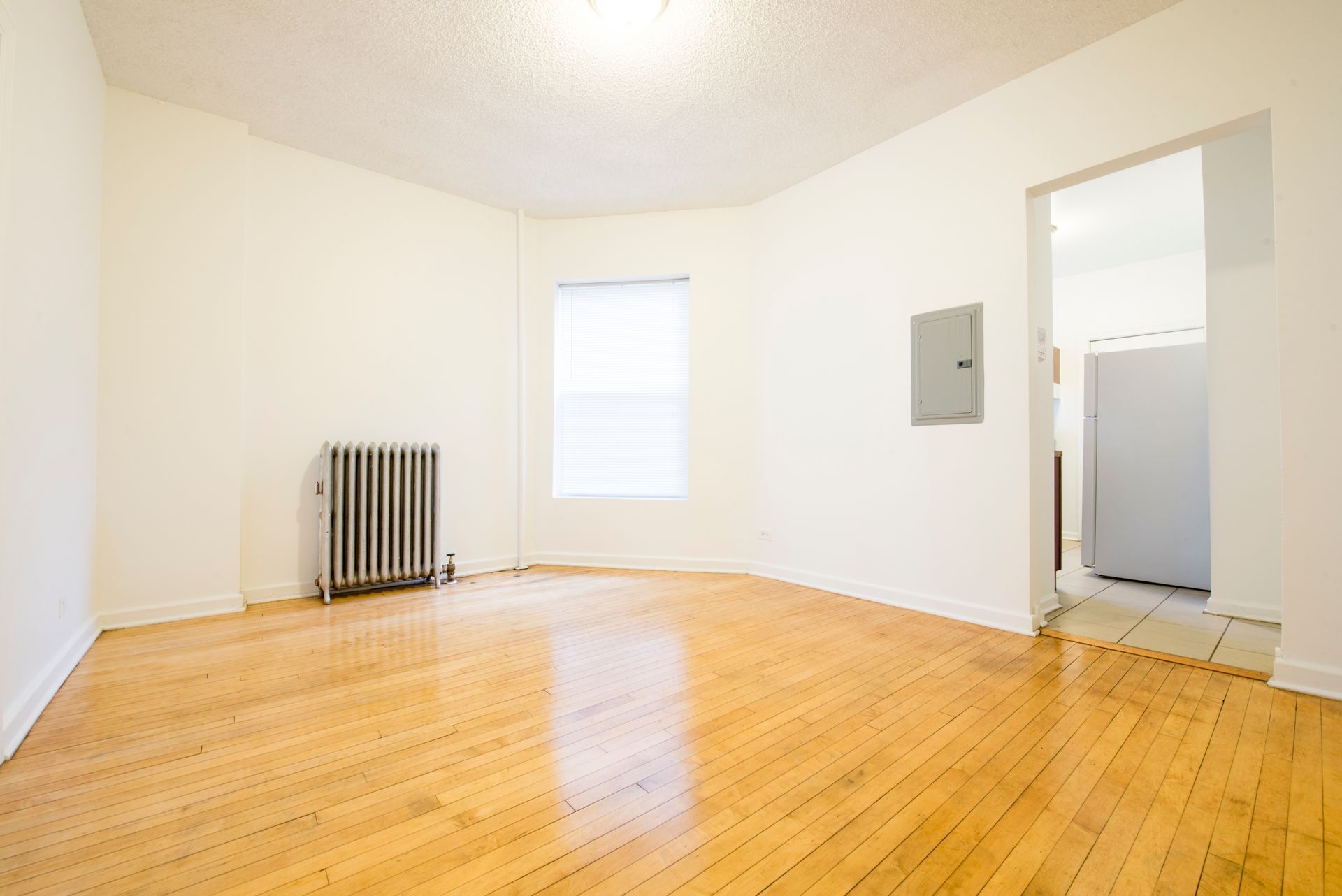 Empty room with hardwood floors, radiator, window, and doorway to a kitchen with a refrigerator.