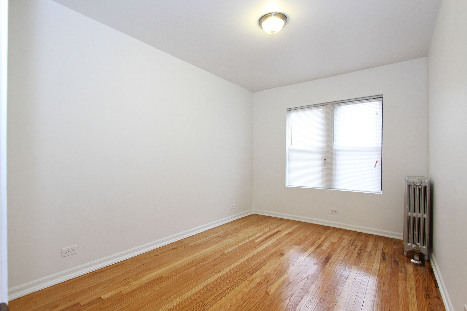 Empty room with hardwood floors, white walls, small window with blinds, and a radiator.