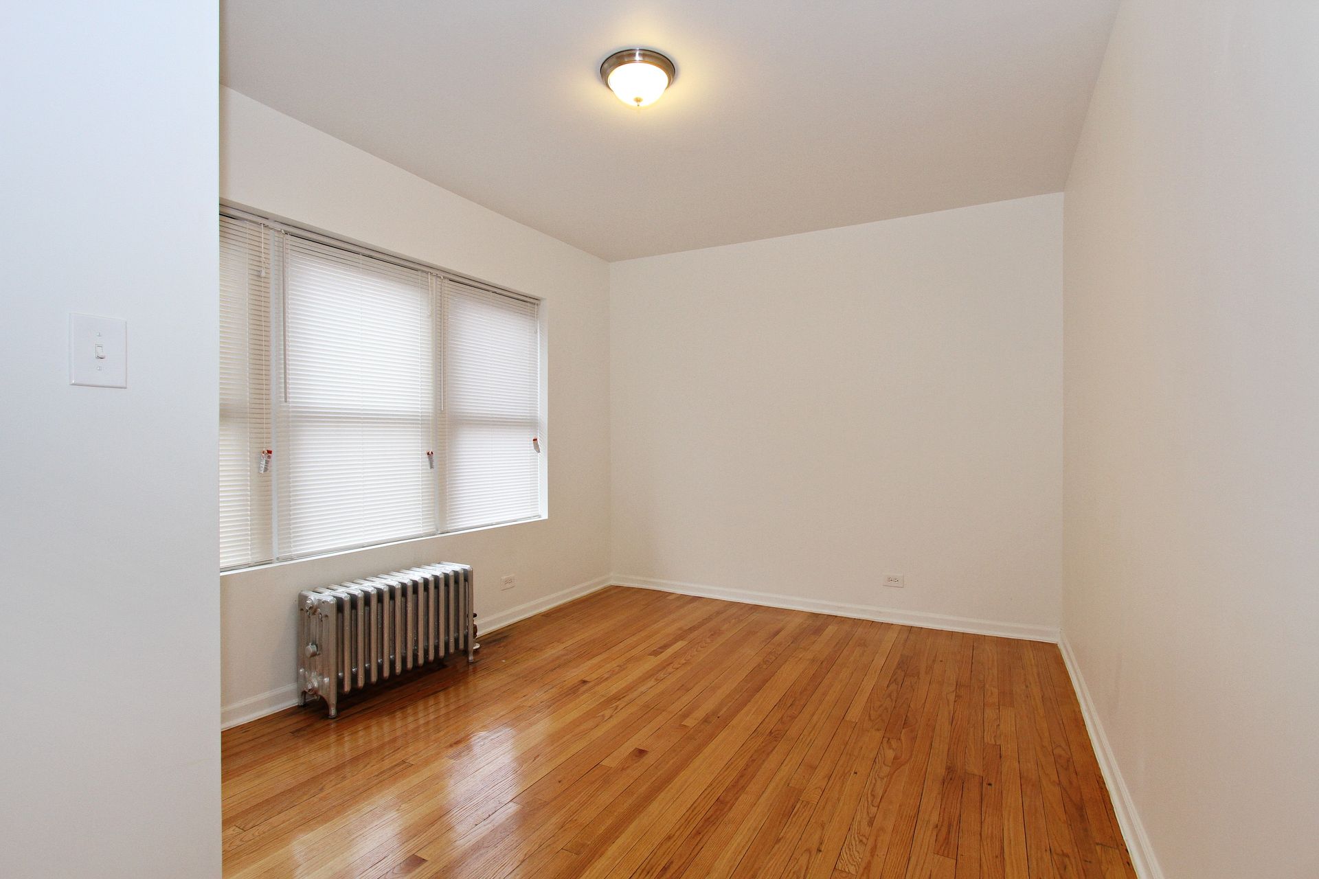 Empty room with hardwood floors, a window with blinds, and a radiator.