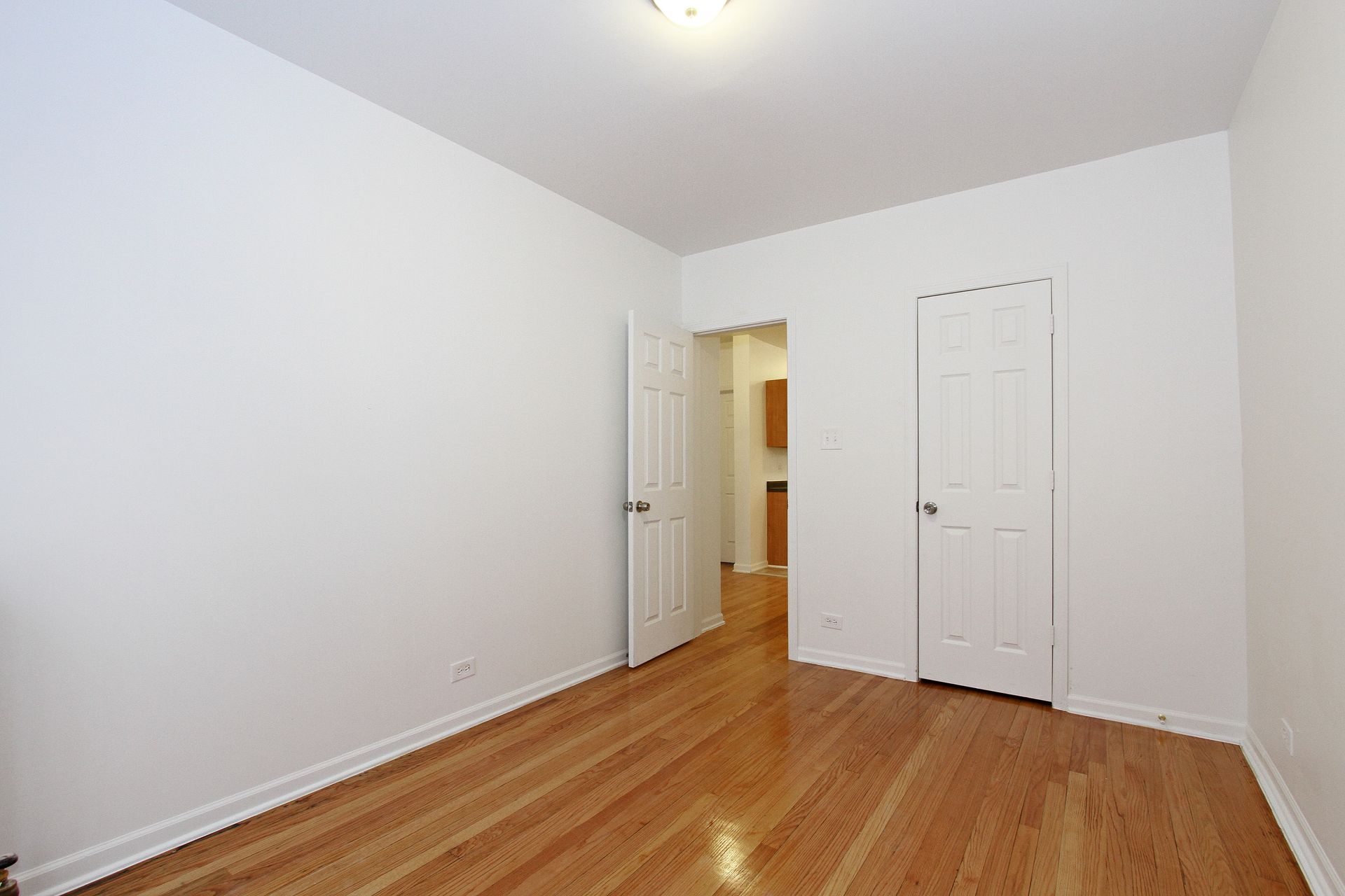 Empty room with hardwood floors, white walls, two white doors, and a ceiling light.