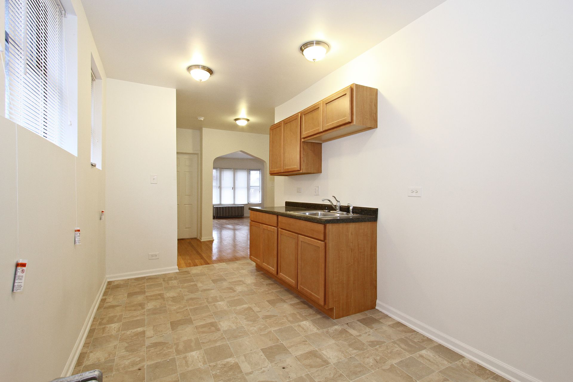 Empty kitchen with wooden cabinets, beige flooring, and a view into another room with hardwood floors.