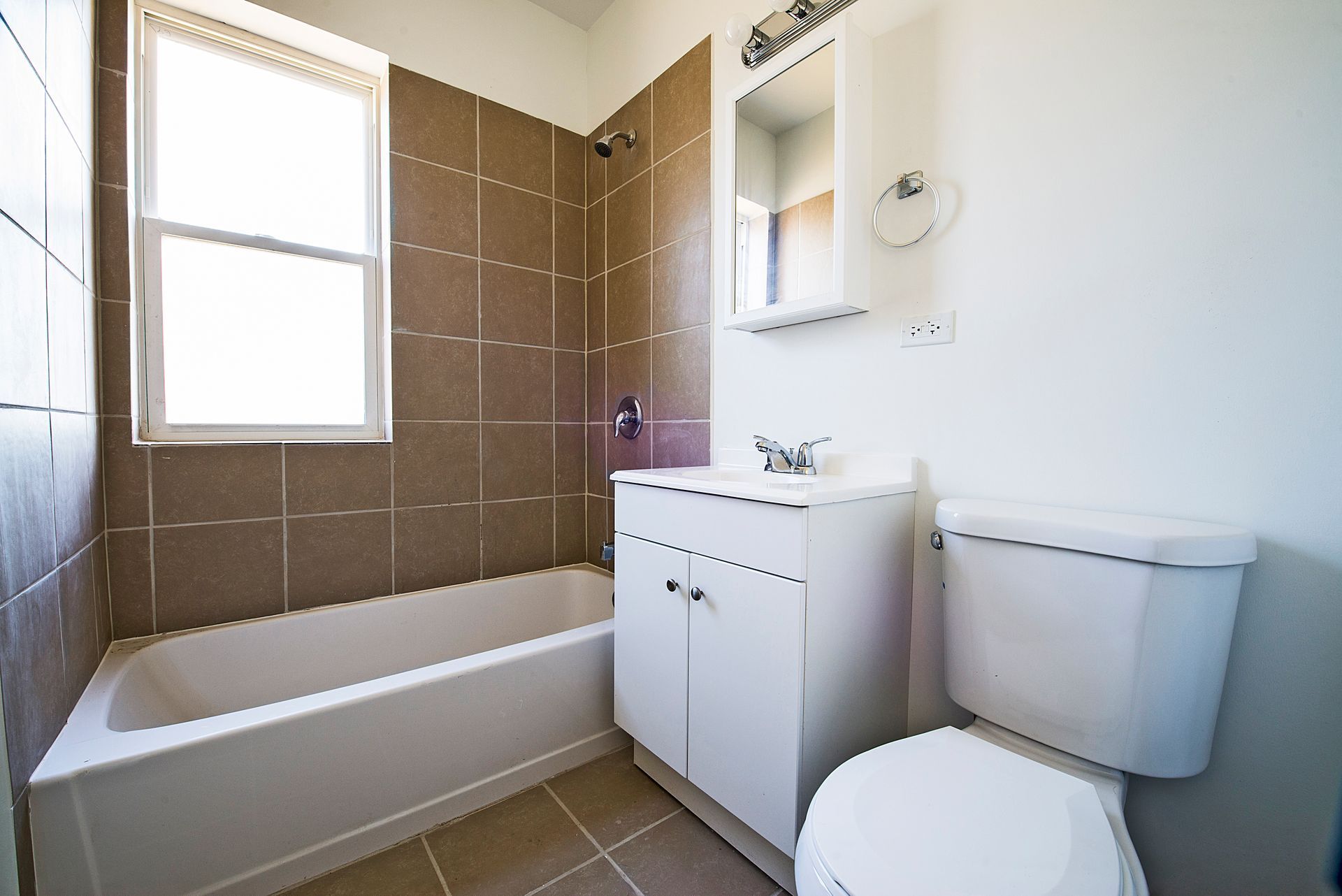 Bathroom with a white bathtub, toilet, and vanity, brown tiled shower and wall, and a window.
