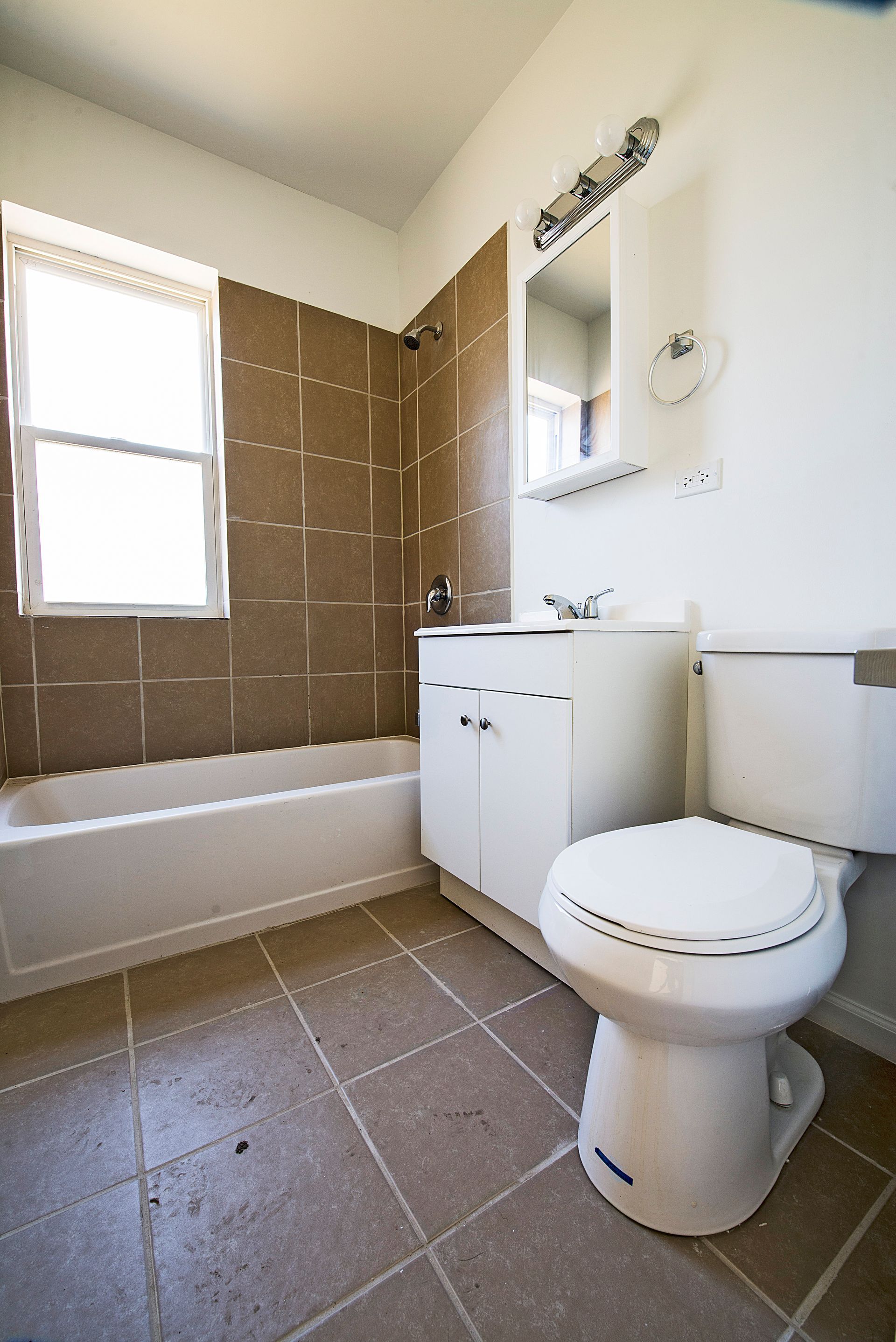 Bathroom with white fixtures, brown tiled wall, tub, and window.
