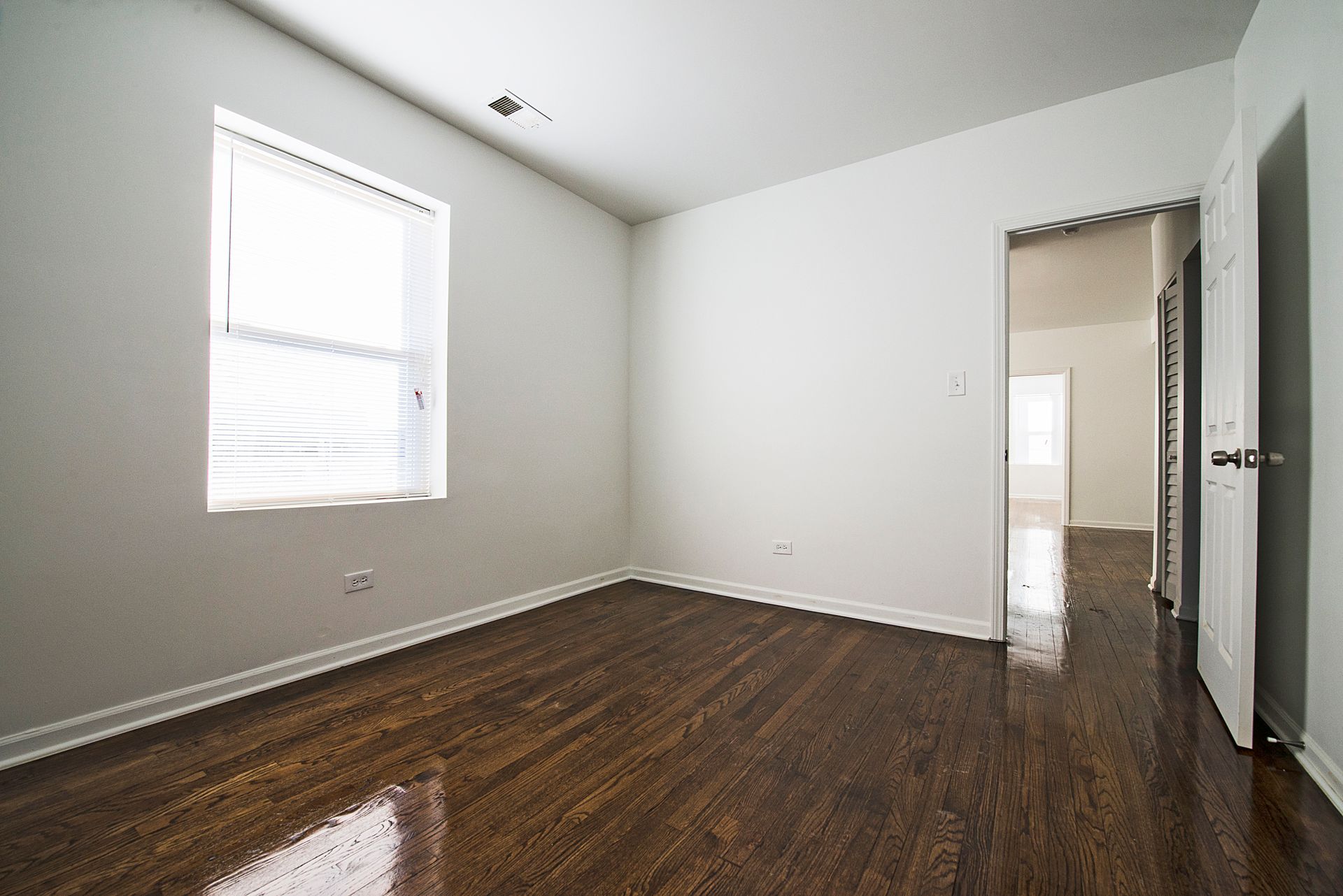 Empty room with dark hardwood floors, white walls, and a window with blinds.
