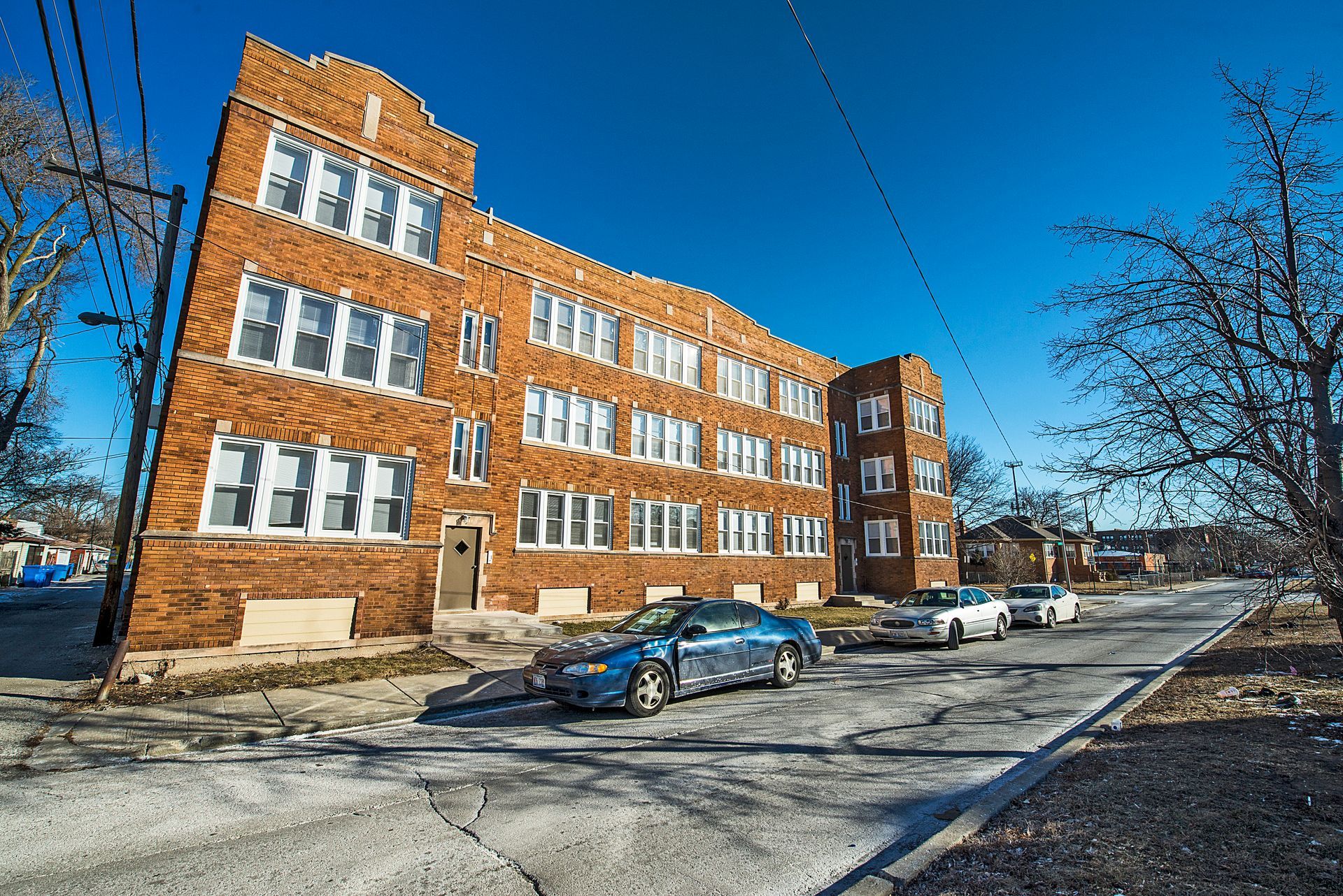 Brick apartment building with parked cars on a cracked street under a clear blue sky.