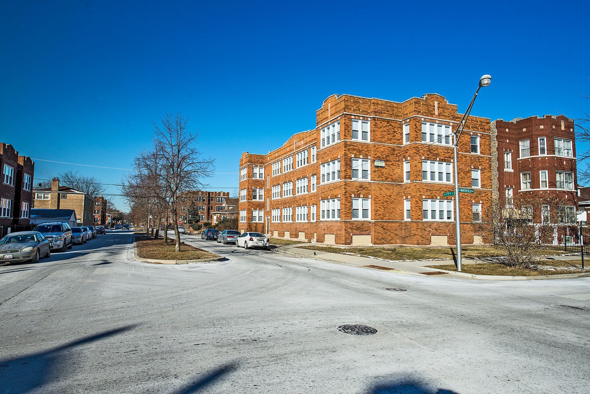 A three-story brick apartment building on a street corner, with parked cars and a clear blue sky.