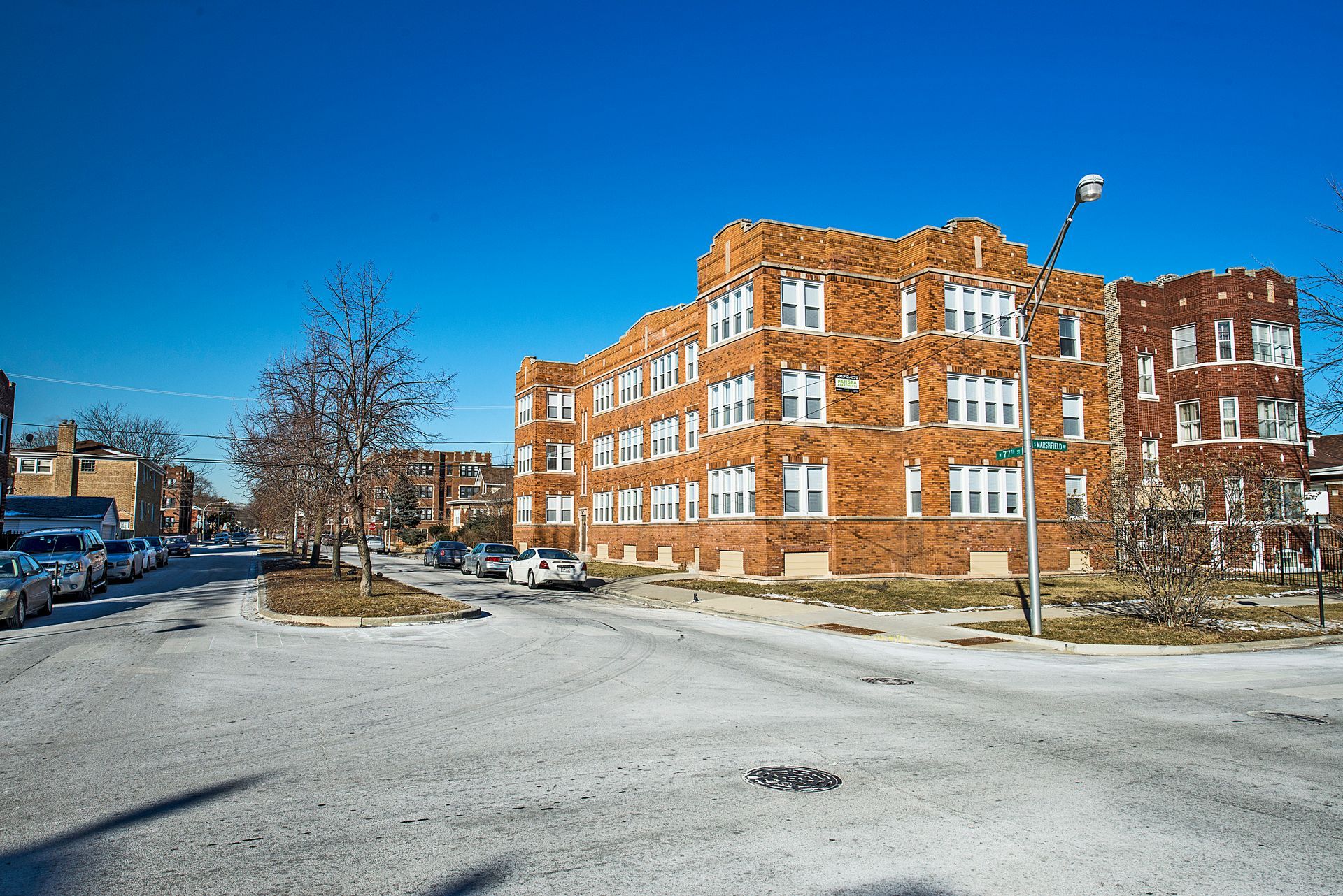 Brick apartment building on a street with bare trees under a clear blue sky.