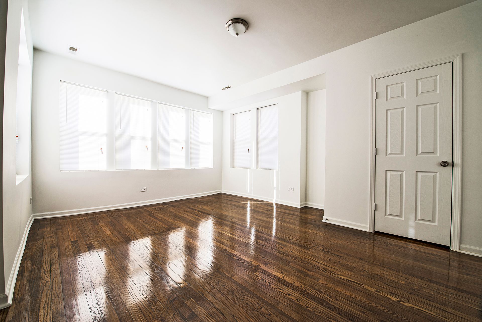 Empty room with dark hardwood floors, white walls, large windows, and a door.