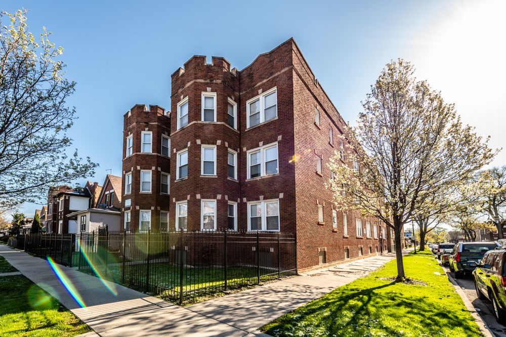 Brick apartment building with turret-like corners, green lawn, sidewalk, and parked cars on a sunny day.