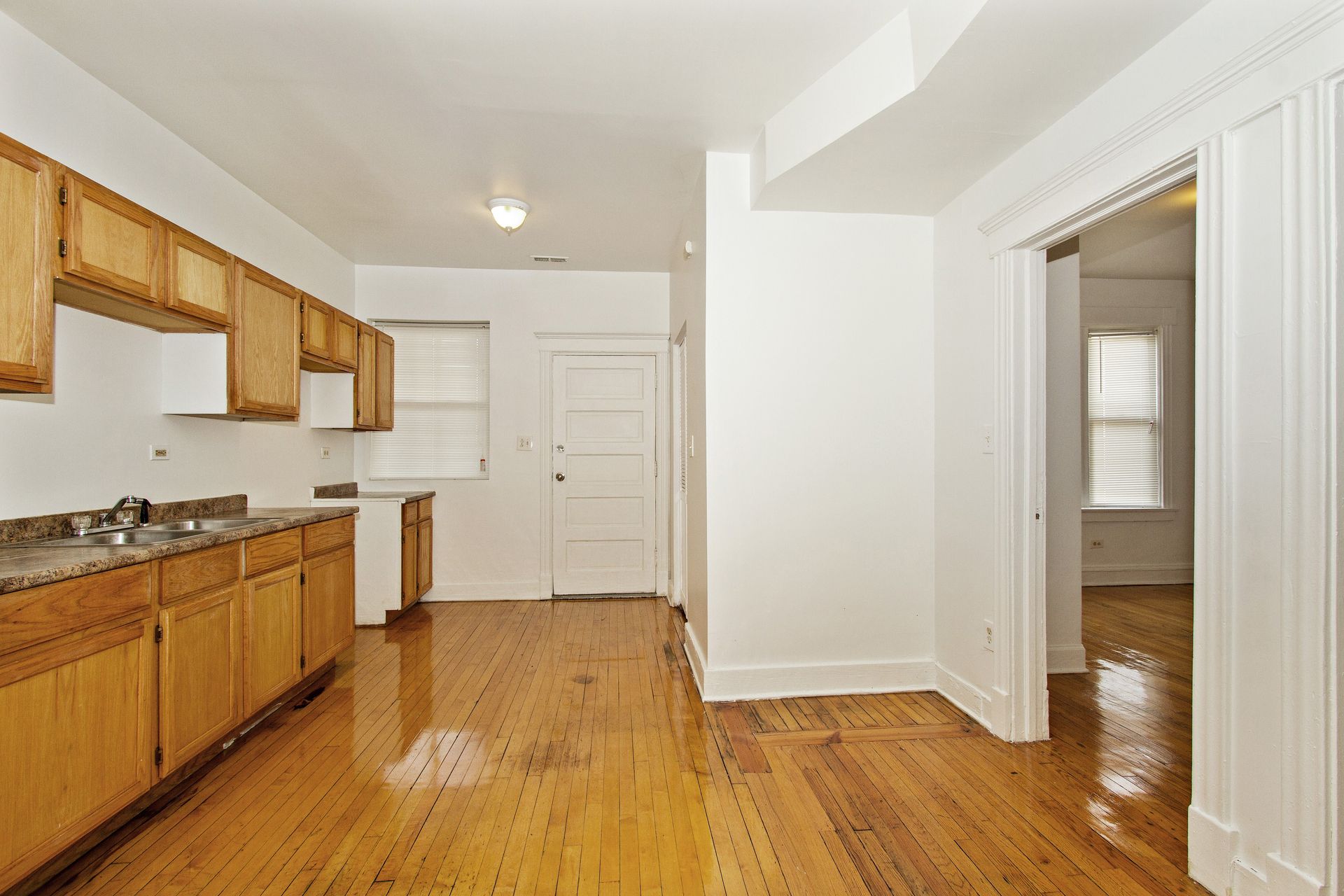 Empty kitchen with wood cabinets, wood floor, and doorway to another room.