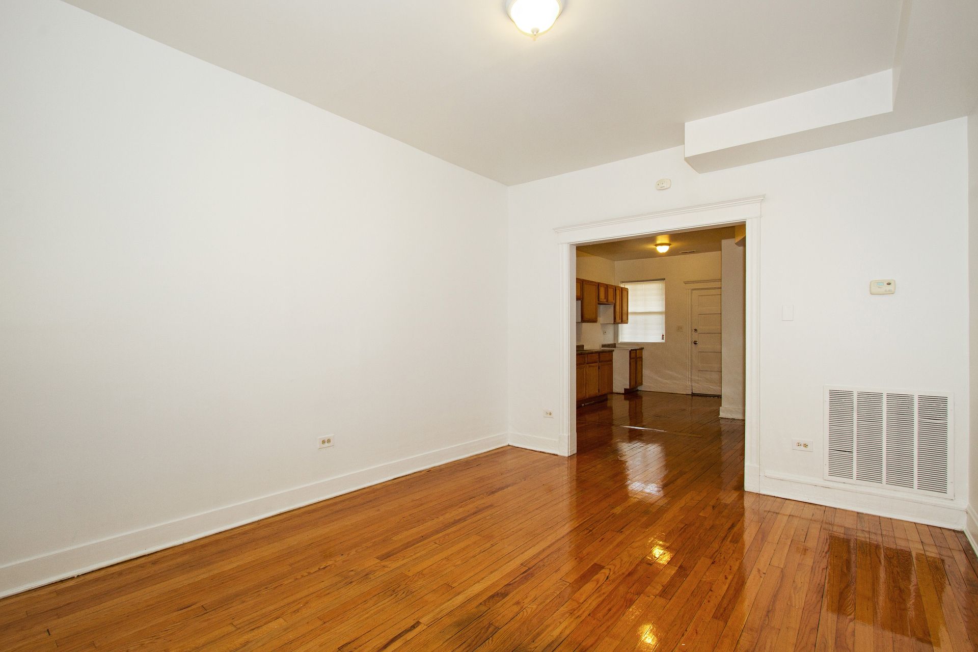 Empty room with hardwood floors, white walls, and an opening to a kitchen.