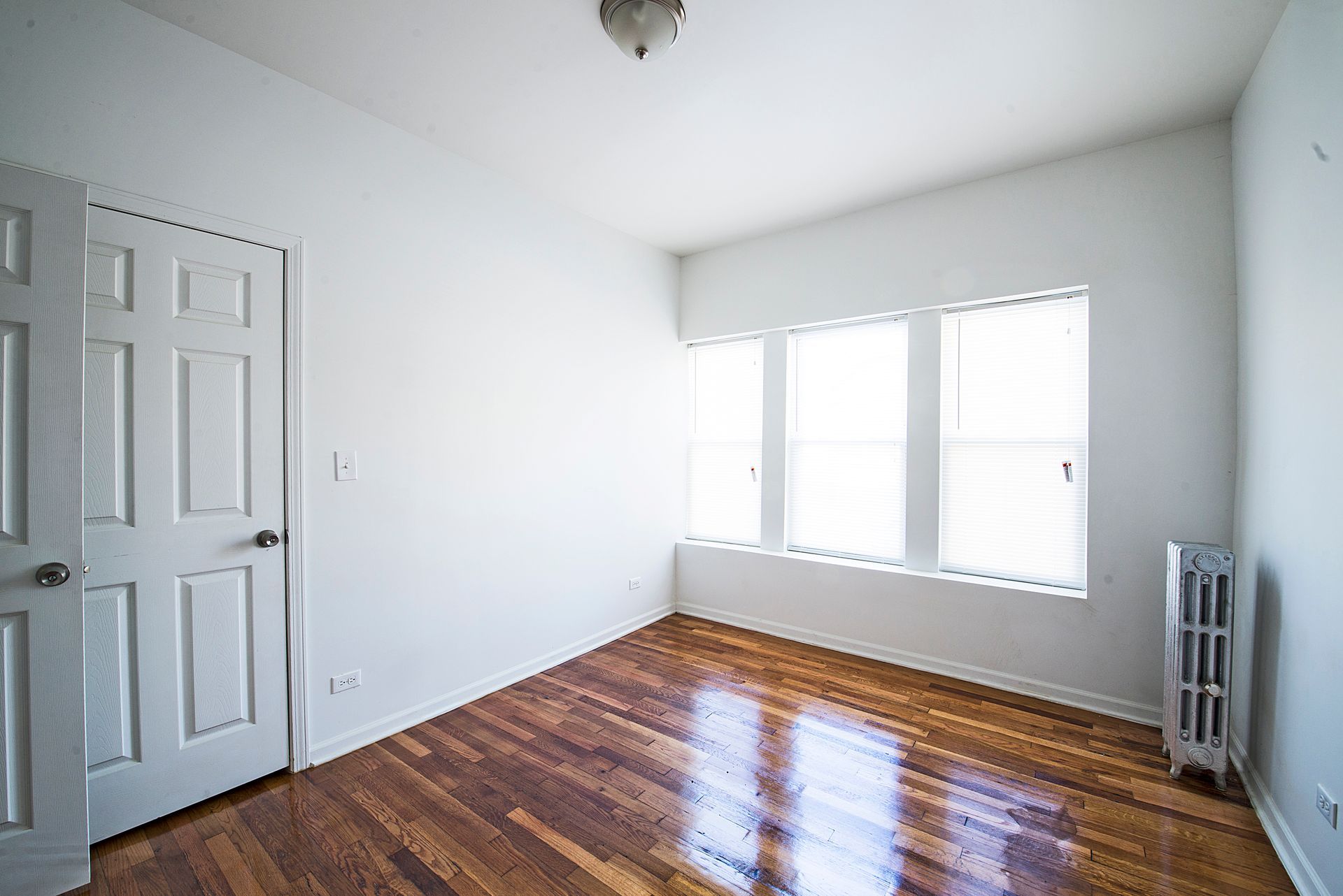 Empty room with wood floor, white walls, door, window, and radiator.