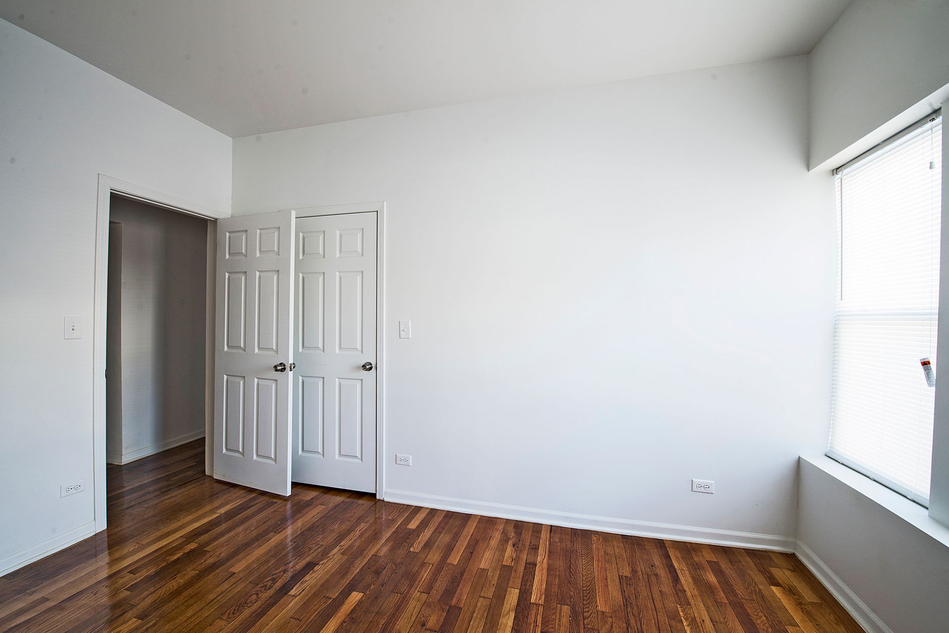 Empty room with hardwood floors, white walls, two closed doors, and a window.