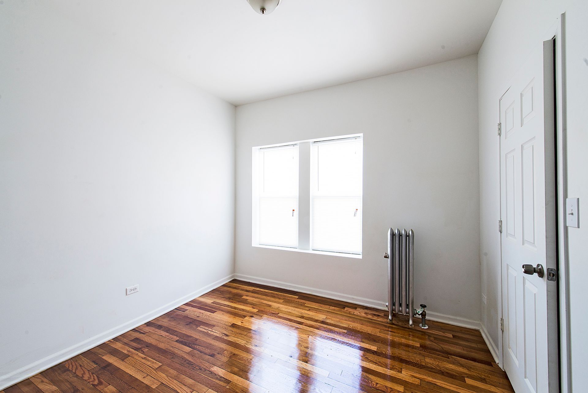 Empty room with hardwood floors, white walls, two windows, radiator, and a closed door.