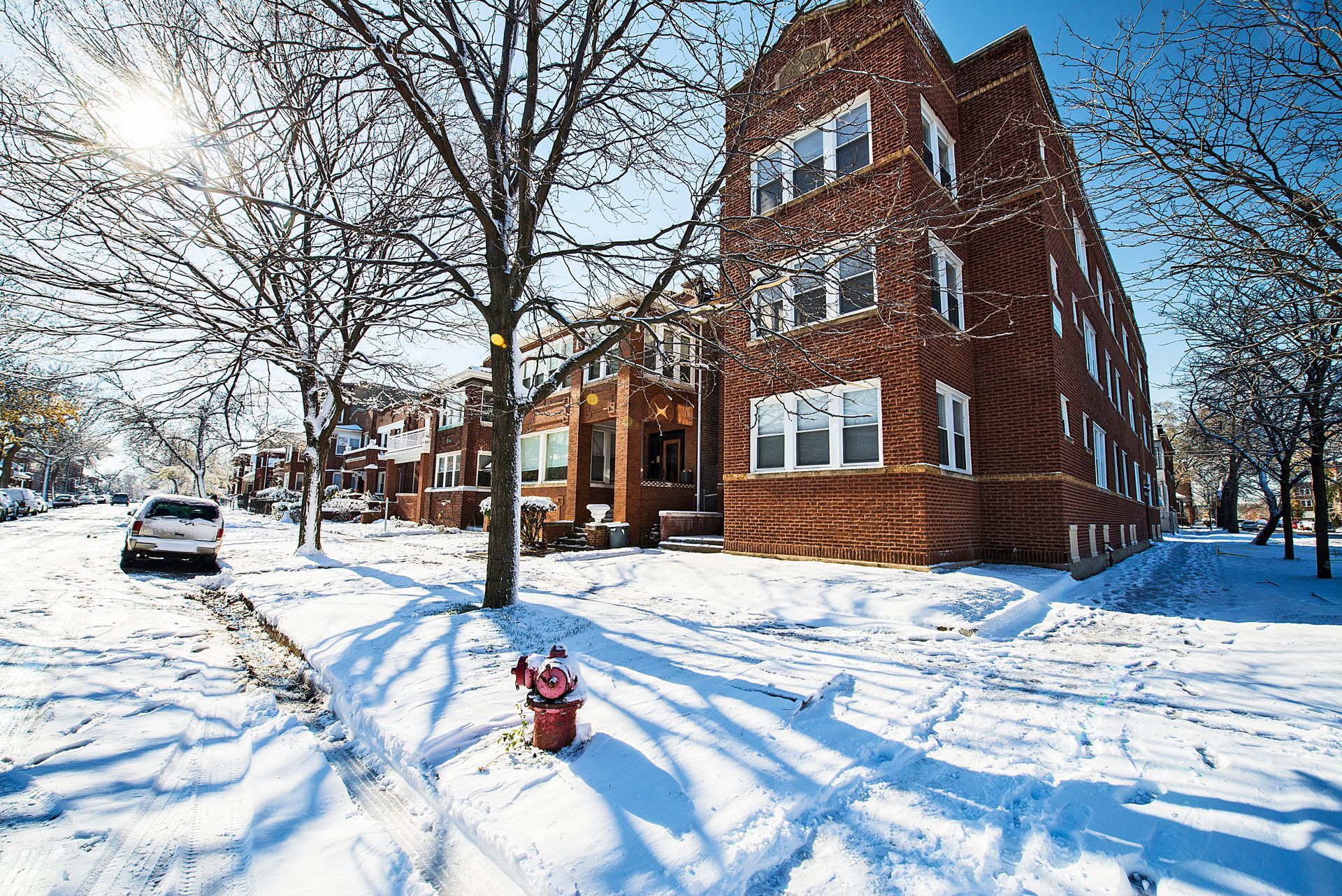 Snowy street scene, brick apartment building, sun shining, parked car, red fire hydrant.
