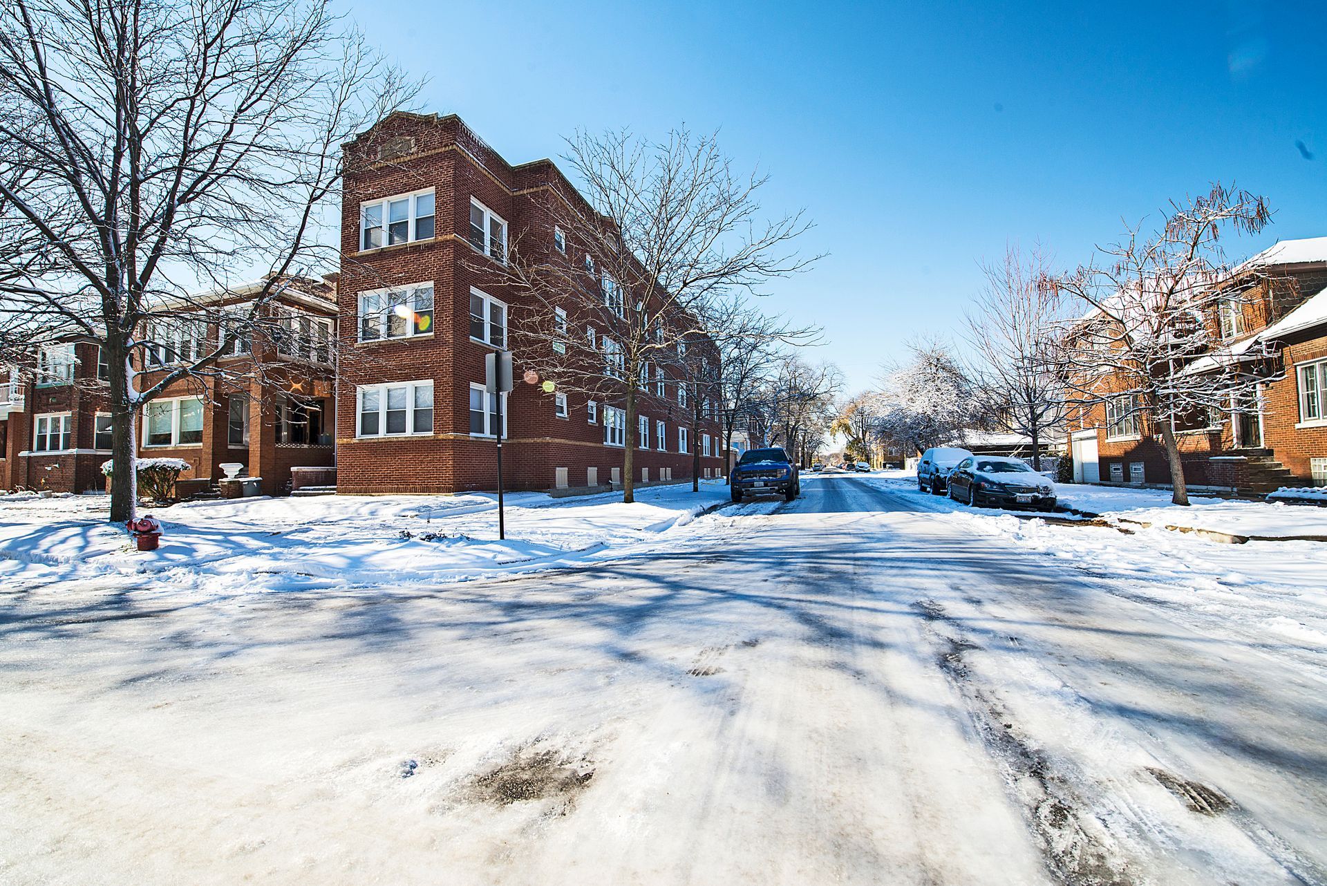 Snowy street with brick apartment building; cars parked along the street.