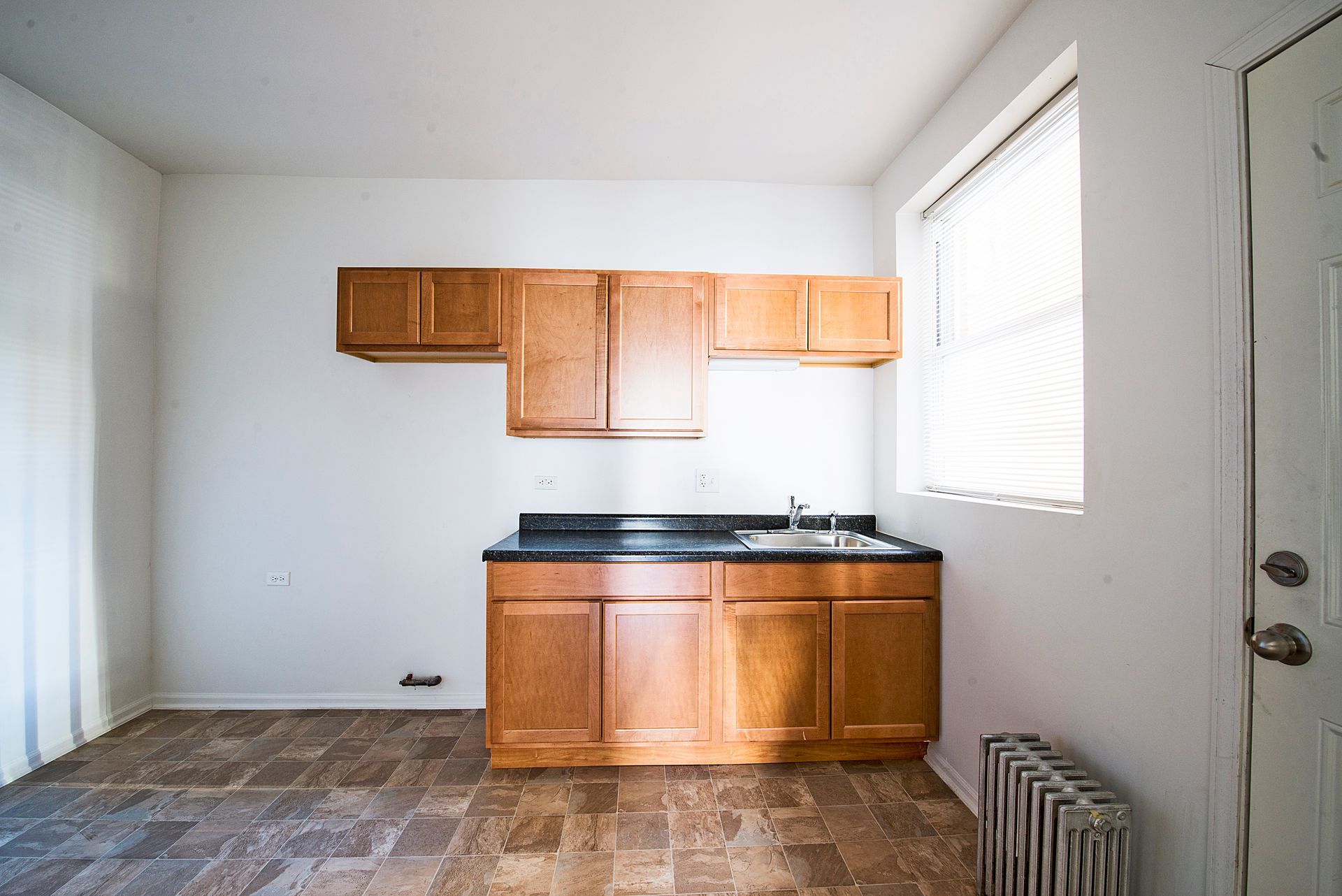 Empty kitchen with wood cabinets, sink, and window; brown flooring, white walls.