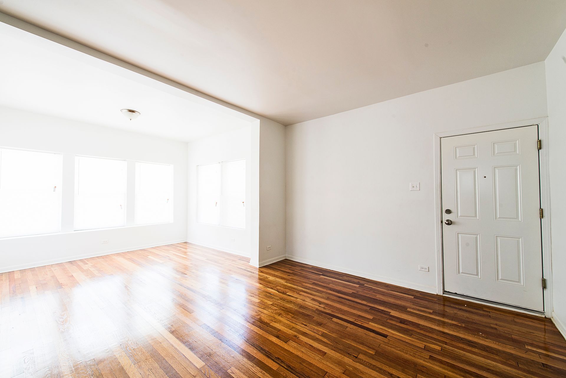 Empty room with hardwood floors, white walls, and a door. Light streams in from the windows.