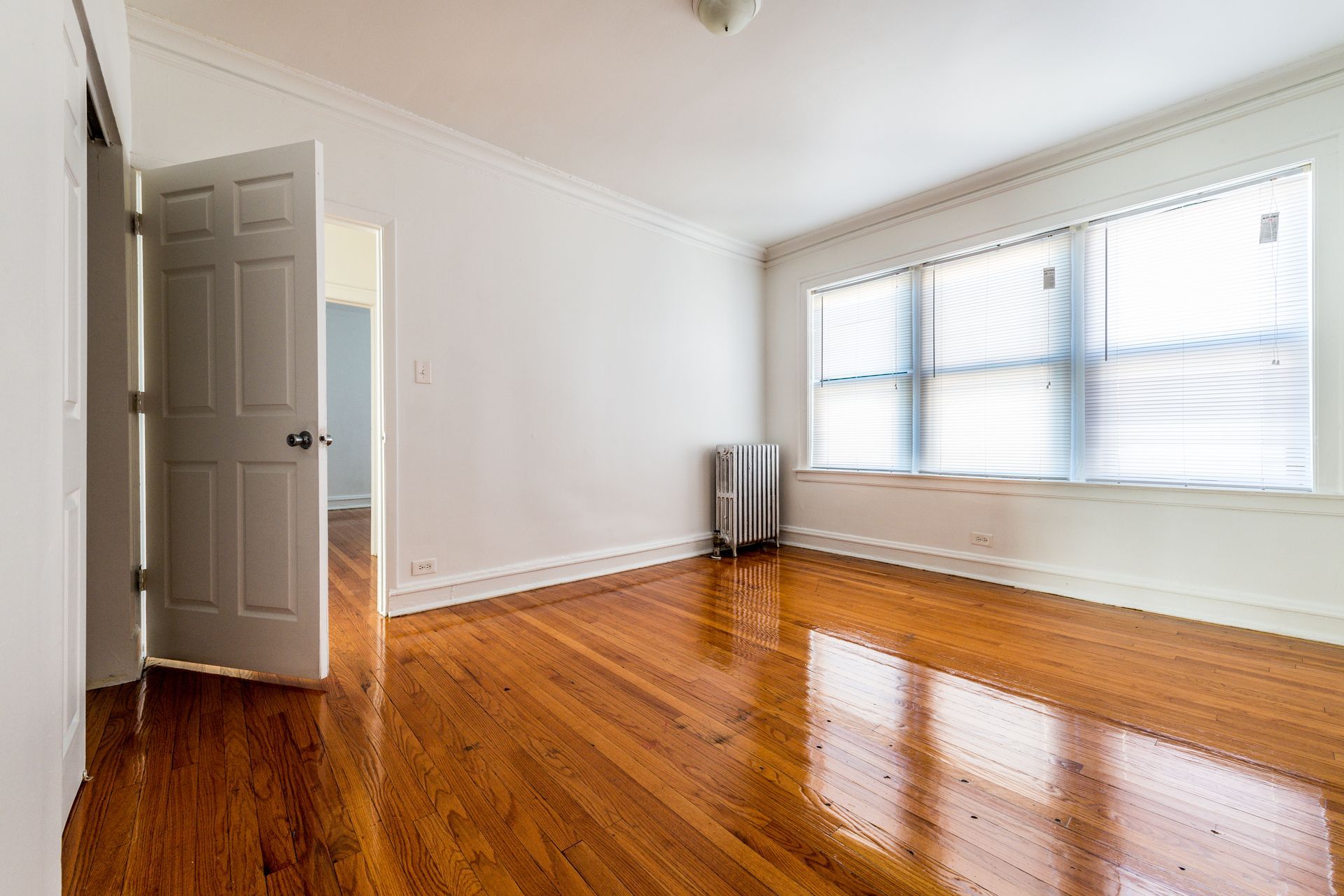 Empty room with wooden floors, white walls, and a window with blinds. Open door to another room.