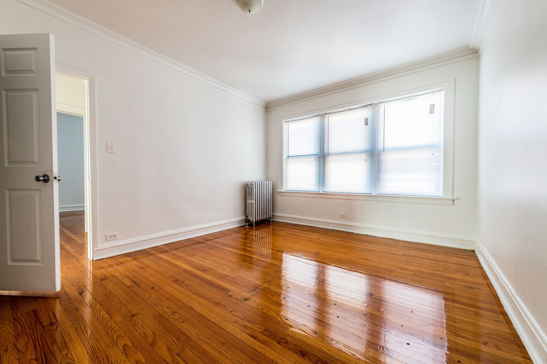Empty room with hardwood floors, a radiator, and a window with blinds. Door open to another room.