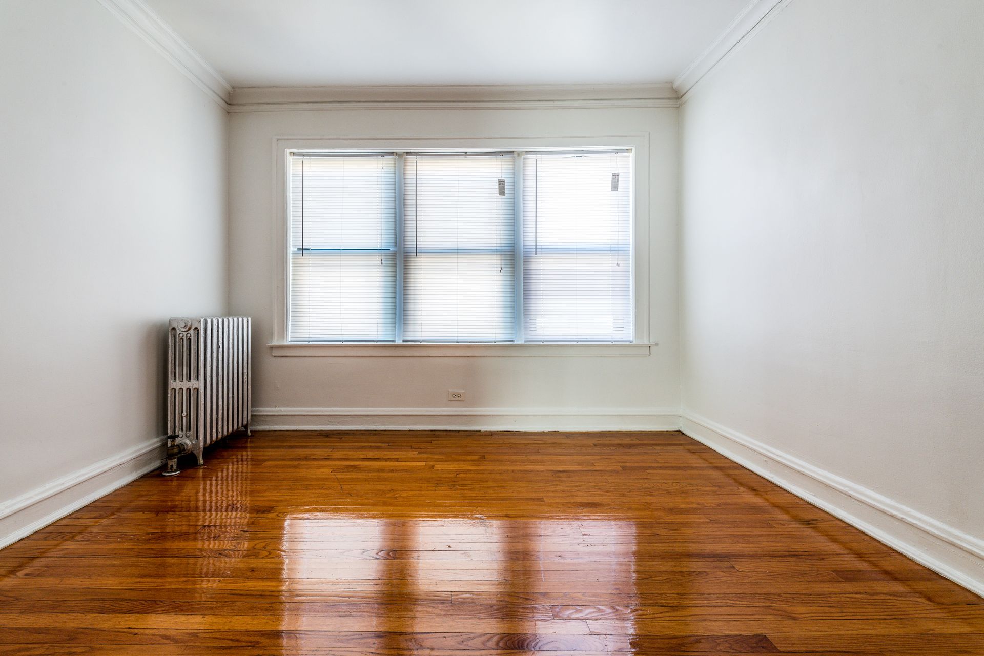 Empty room with wood floor, white walls, radiator, and window with blinds.