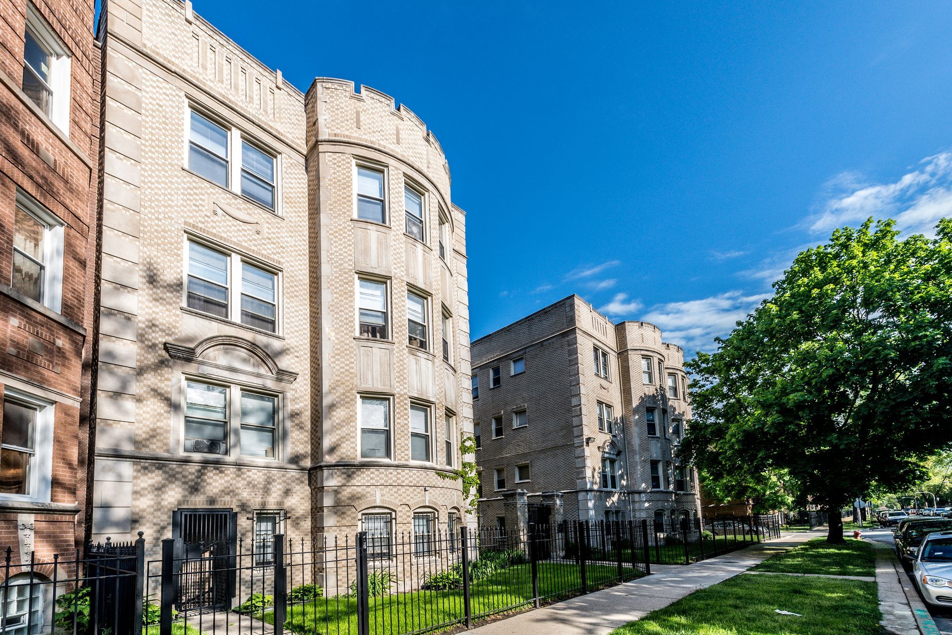 Brick apartment buildings with arched details, sidewalk, trees, and blue sky.