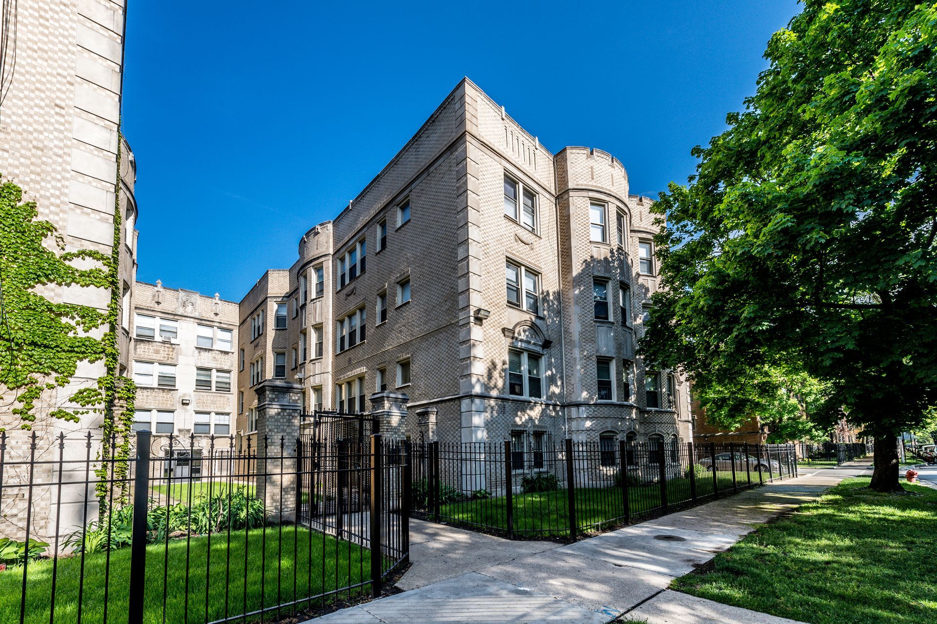 Apartment building with light brick facade, green lawn, iron fence, and large tree under a bright blue sky.
