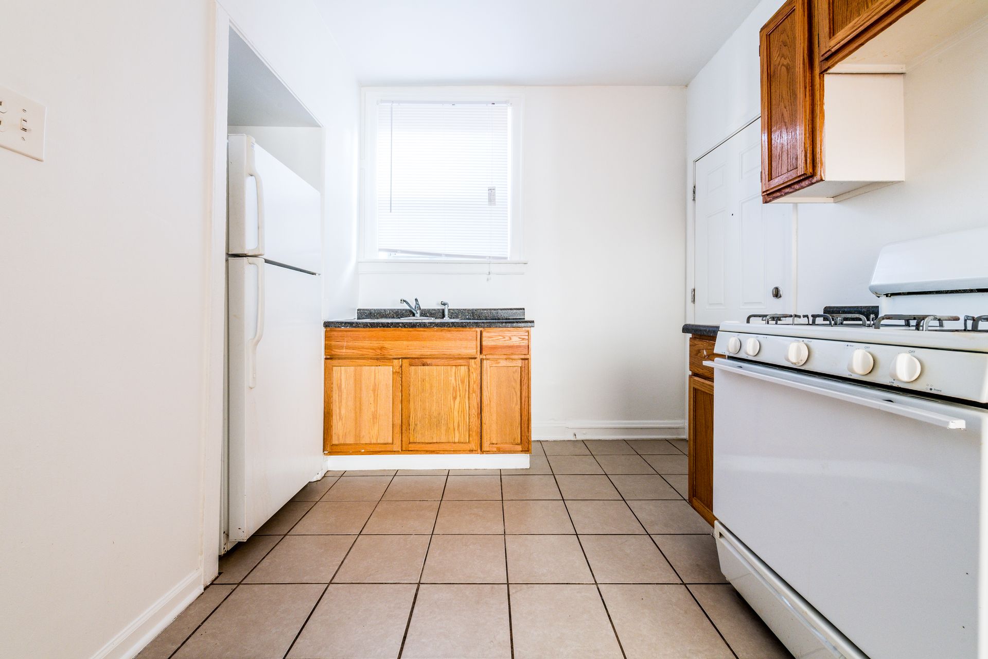 Kitchen with wood cabinets, white appliances, window above sink, and tile floor.