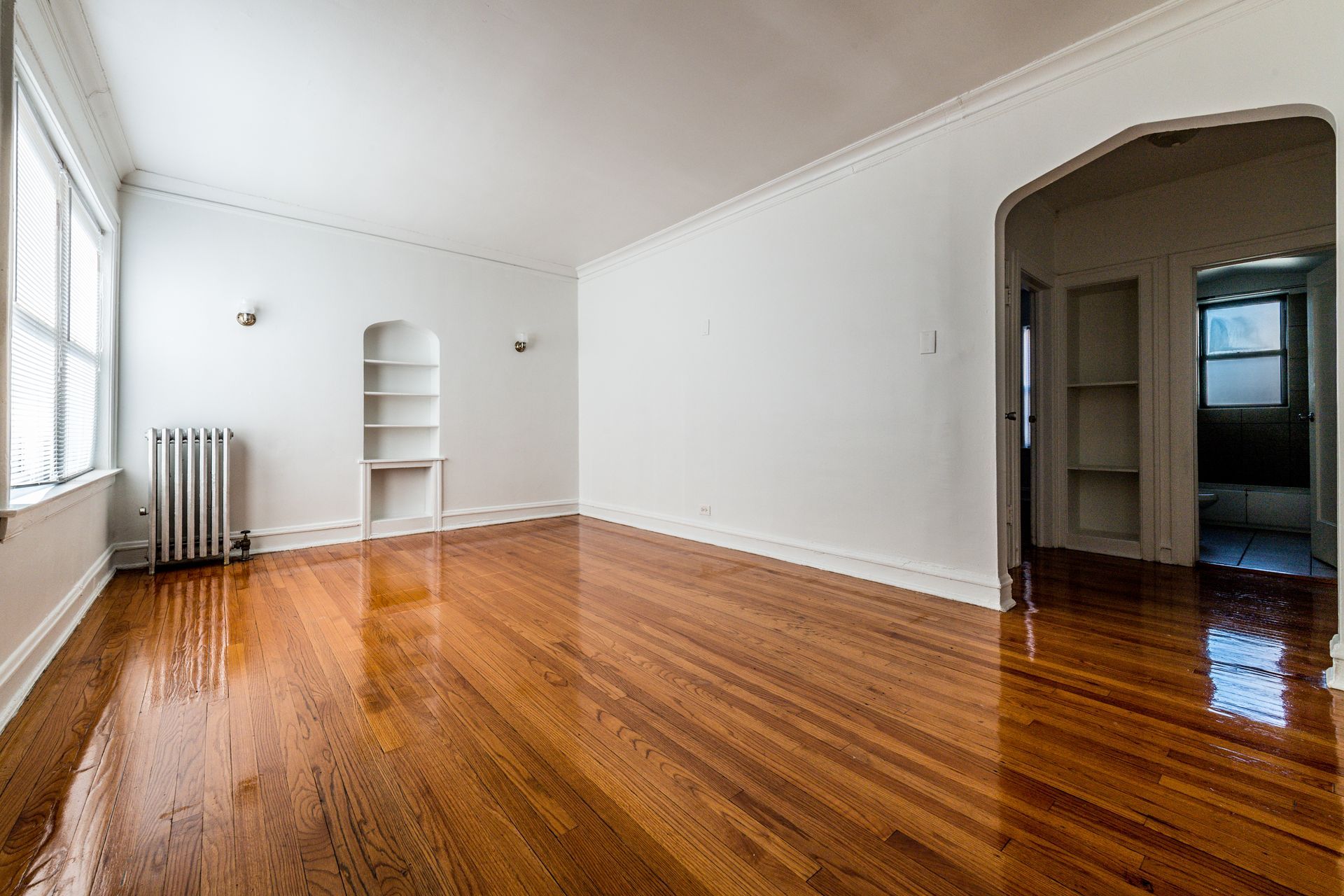 Empty living room with wood floors, radiator, built-in shelving, and archway to other rooms.