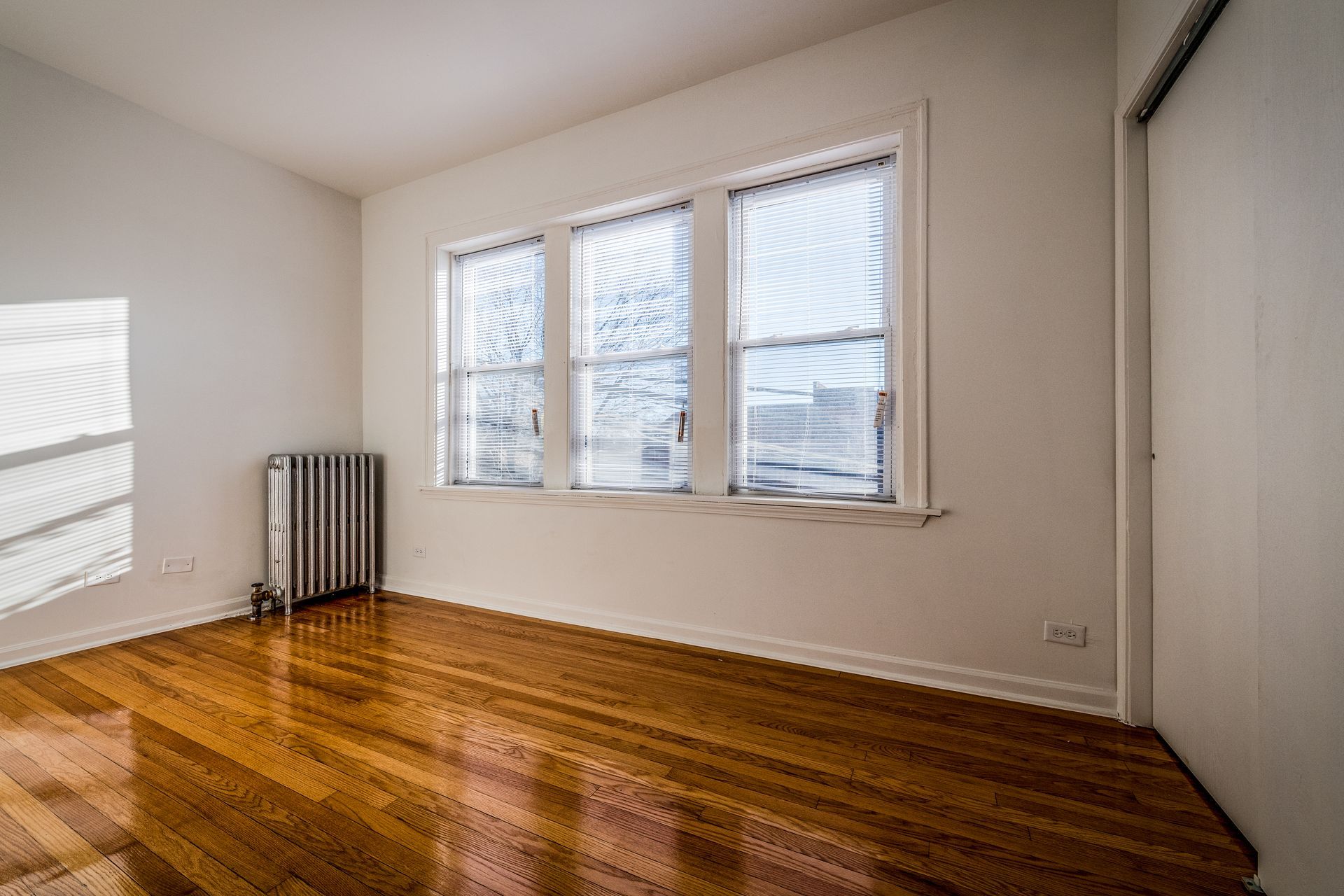 Empty room with hardwood floor, three windows, radiator, and a white closet door.