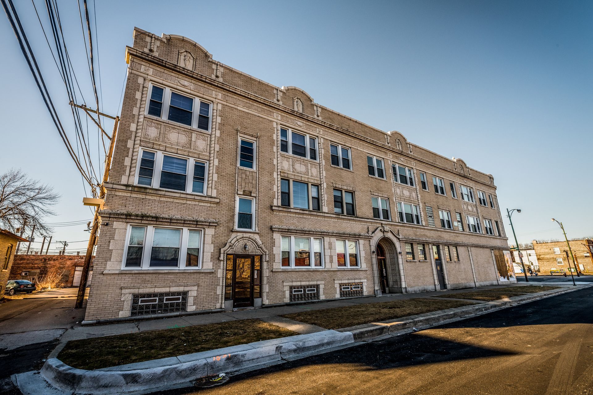 Three-story brick apartment building on a street, with windows and a decorative top, on a sunny day.