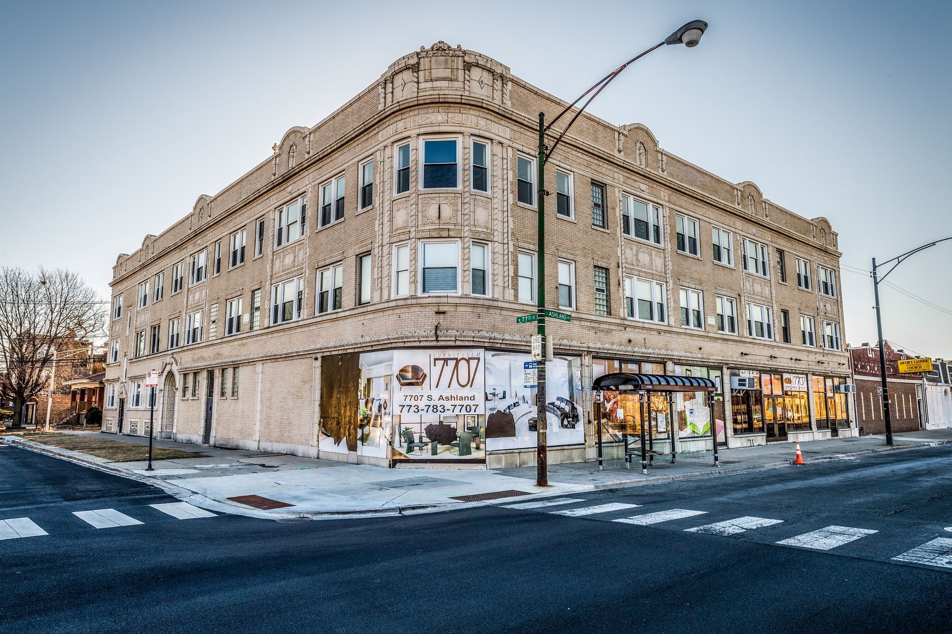 A three-story, beige brick building on a street corner, with storefronts and windows.