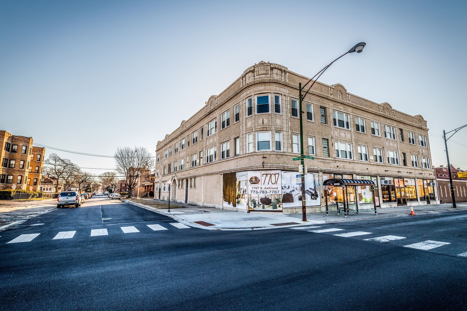 Corner building, two stories, storefronts, brick facade. Empty street, crosswalk. Cloudy sky.